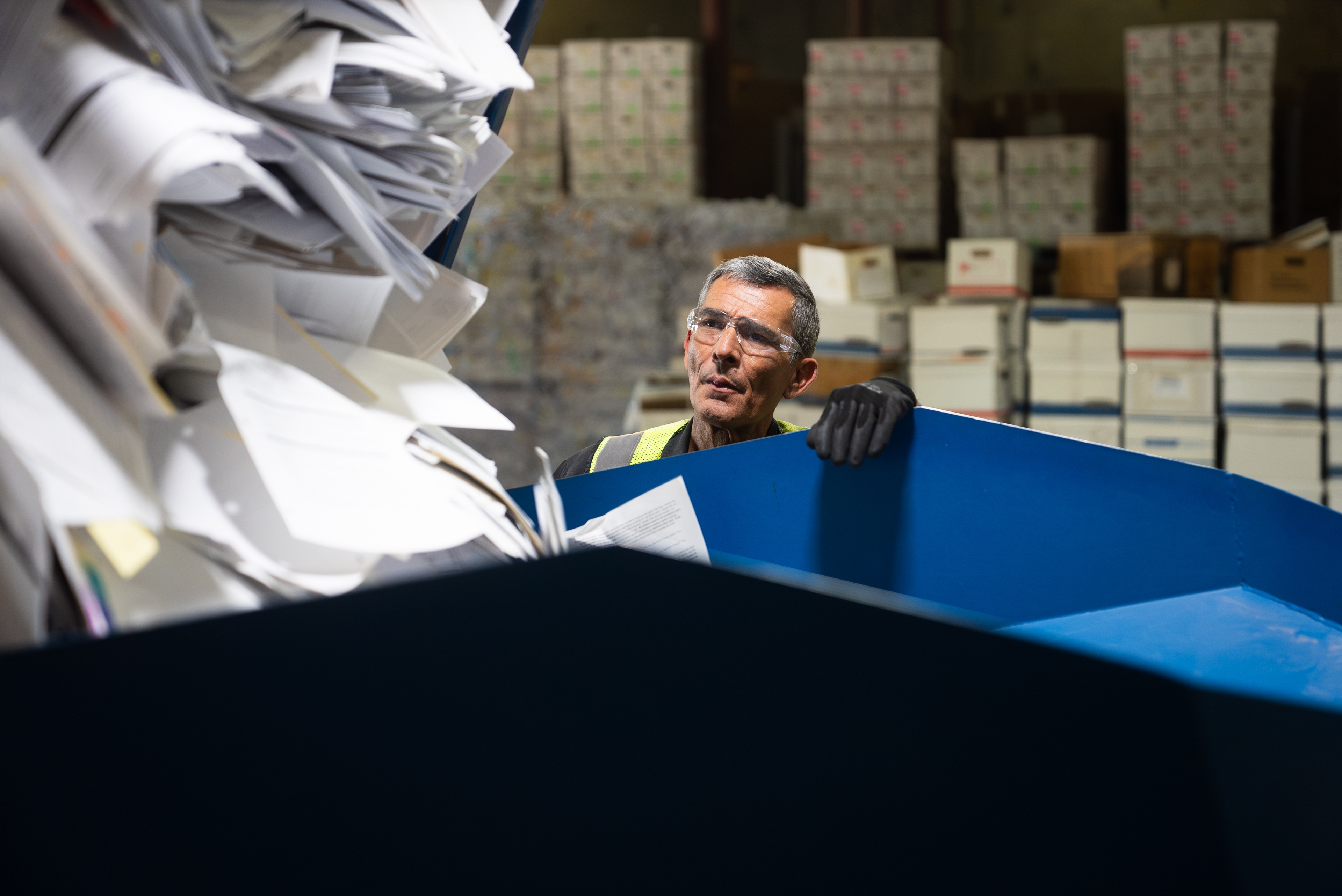 Warehouse worker loading loose paper documents into a secure shredding container at a document destruction facility, with records storage boxes in the background.