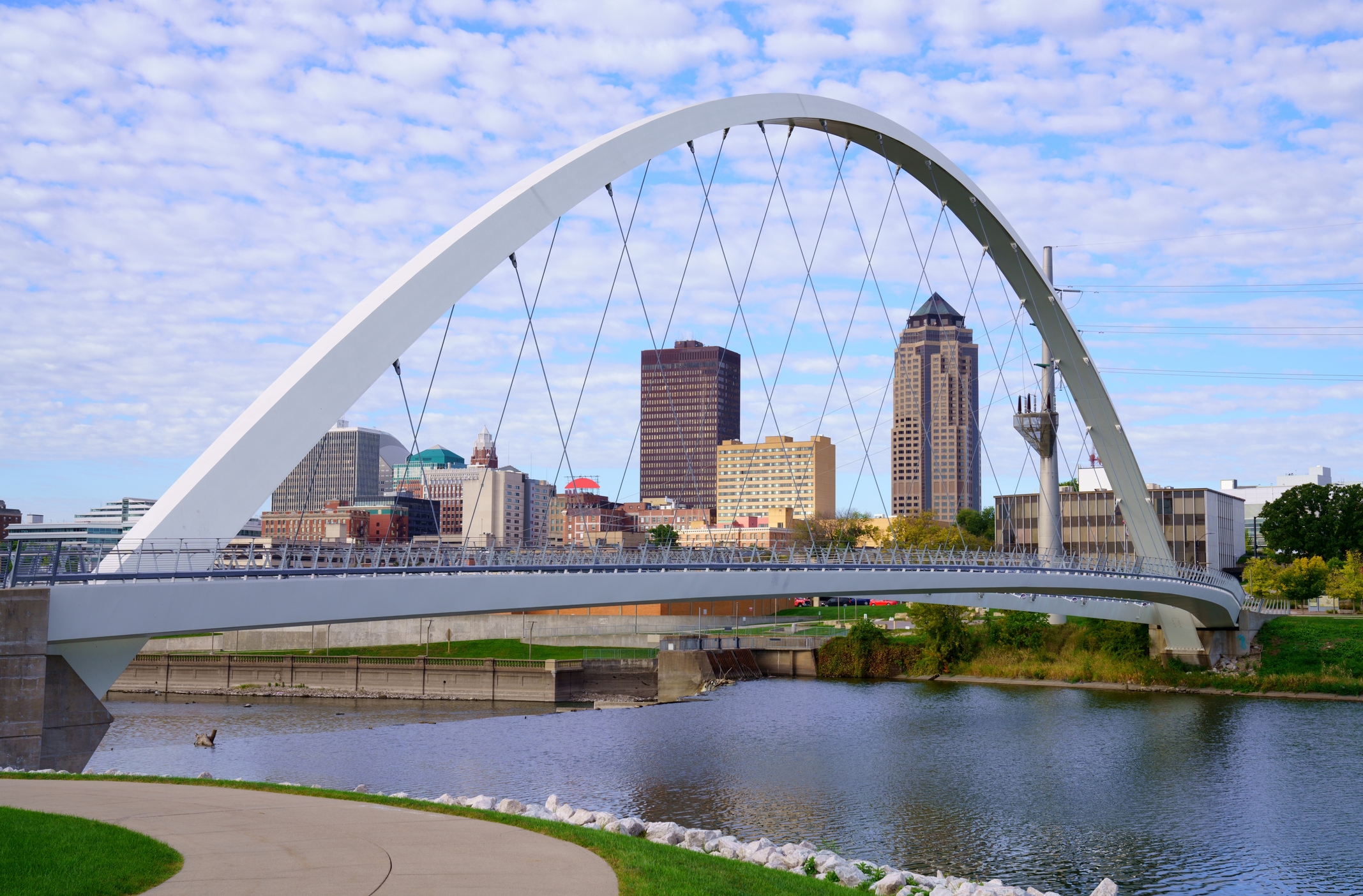 A view of Des Moines, Iowa, featuring the iconic arching pedestrian bridge spanning a calm river, with the city skyline, including tall buildings, in the background under a partly cloudy sky.