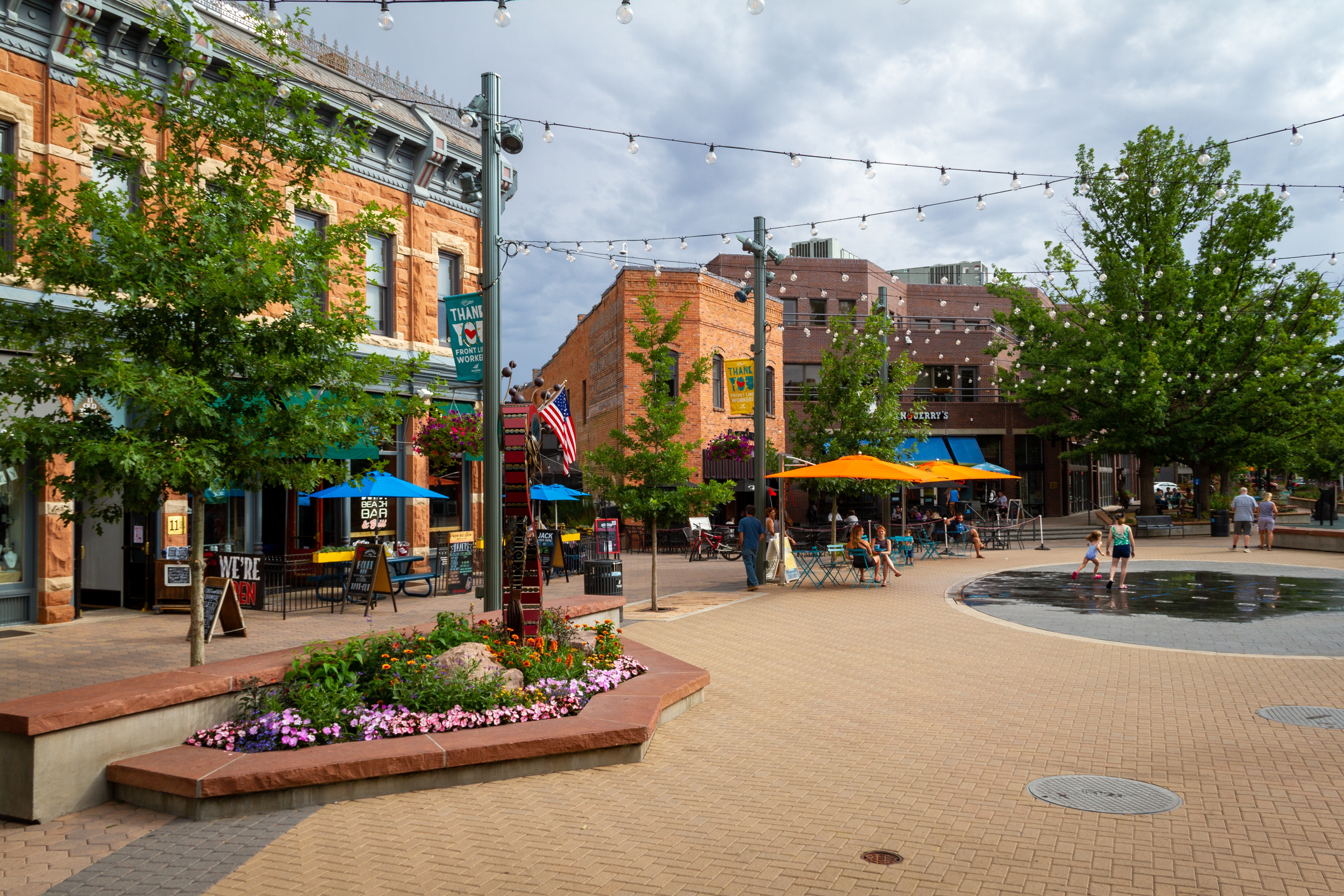 Downtown Fort Collins, Colorado, featuring historic brick buildings, colorful umbrellas over outdoor seating, a splash pad with children playing, string lights overhead, and vibrant flower beds on a sunny afternoon.