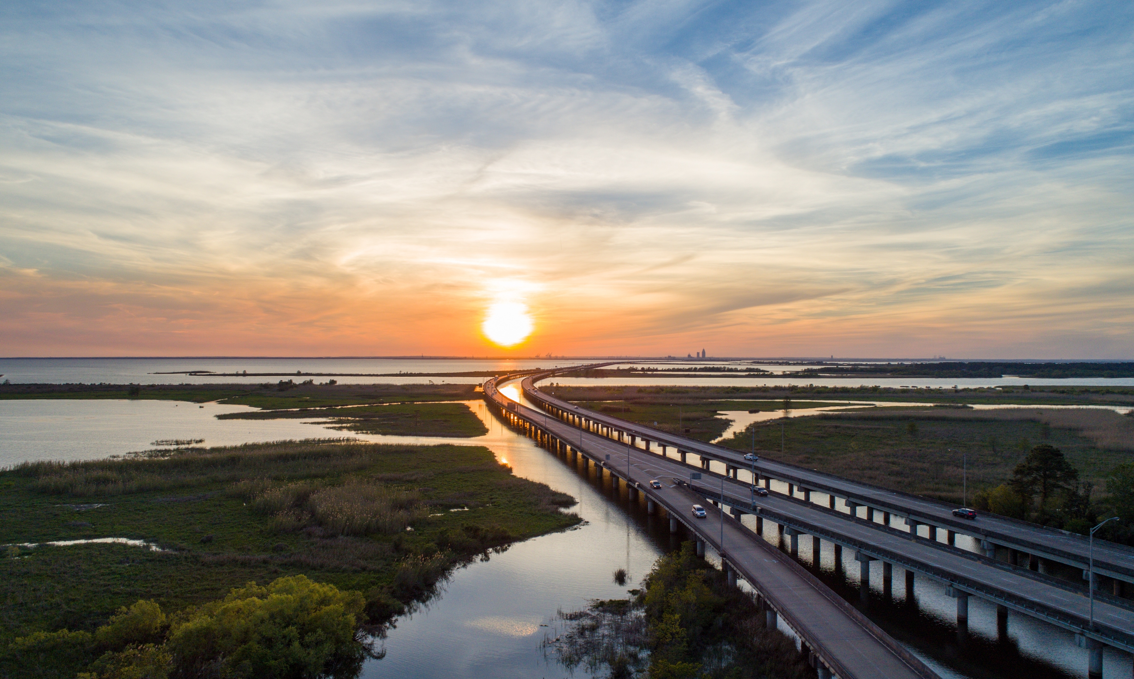 Scenic aerial view of the Mobile Bay Causeway at sunset, with traffic crossing over reflective waters and the distant Mobile skyline silhouetted against a glowing Gulf Coast horizon.