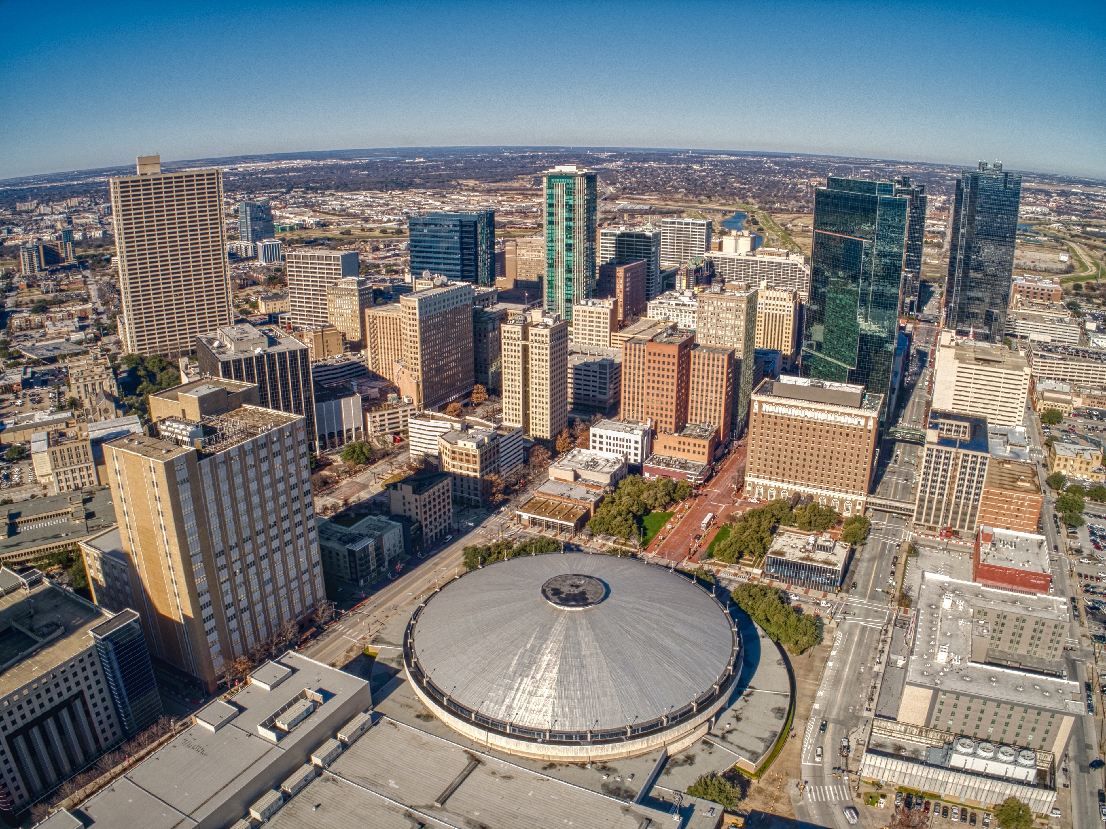 Aerial view of downtown Fort Worth, Texas, showcasing a dynamic mix of high-rise buildings, historic architecture, and the iconic circular Fort Worth Convention Center under a clear blue sky.

