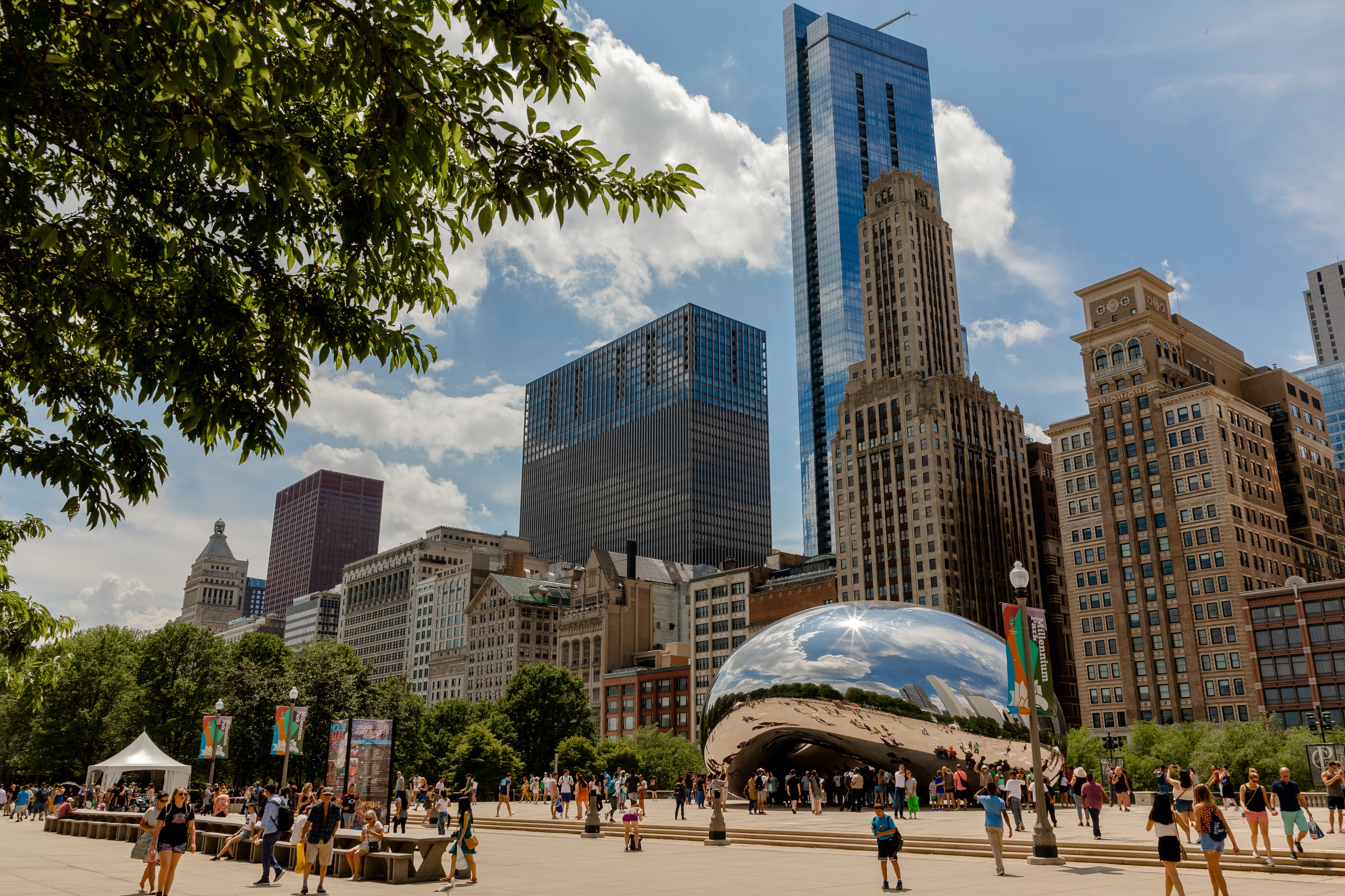 View of Cloud Gate (“The Bean”) sculpture in Chicago’s Millennium Park with crowds of people and historic and modern skyscrapers in the background on a sunny day.