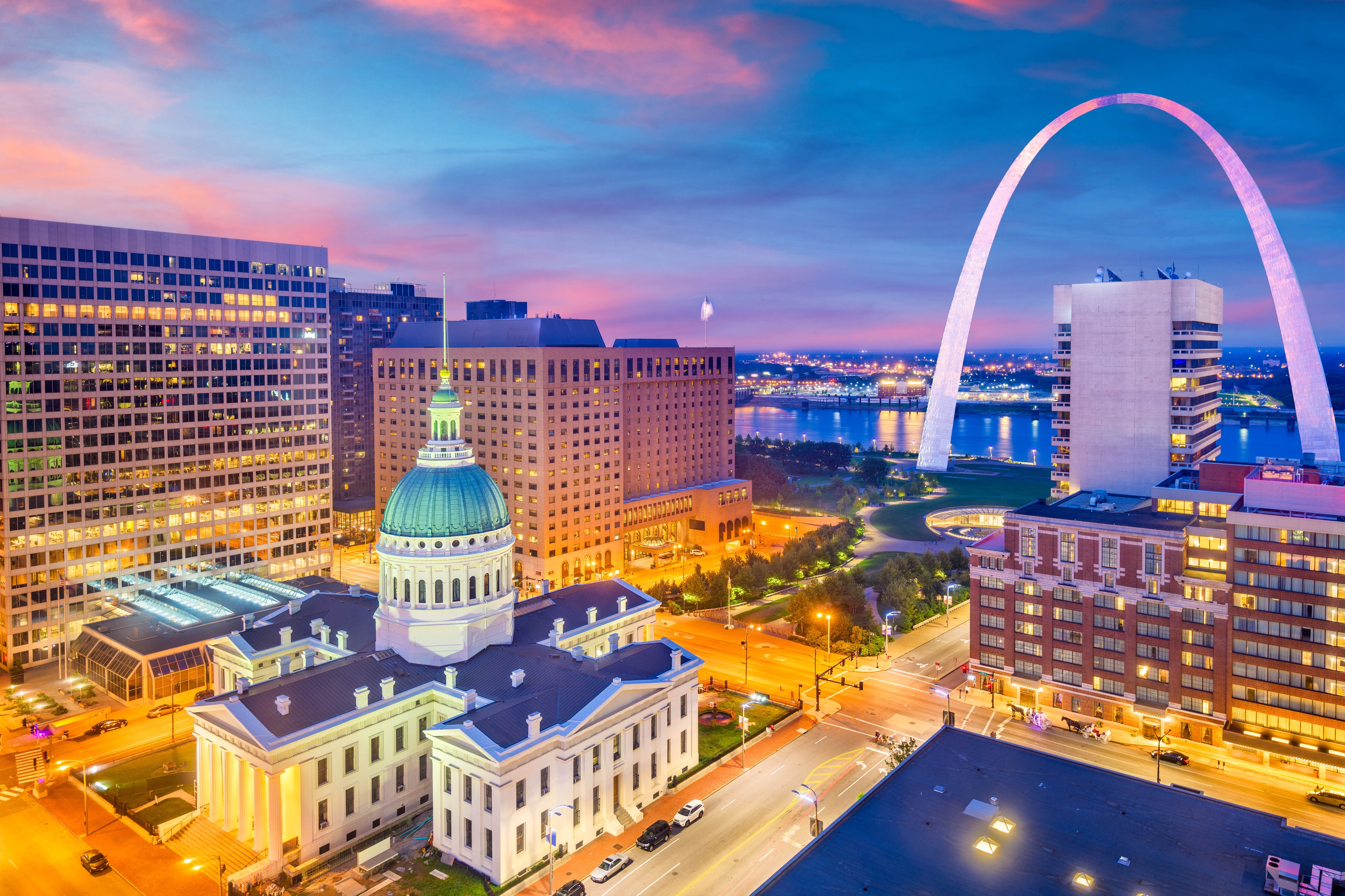 A vibrant twilight view of downtown St. Louis featuring the Old Courthouse with its green dome, the Gateway Arch illuminated in purple light, and the Mississippi River glowing in the background.