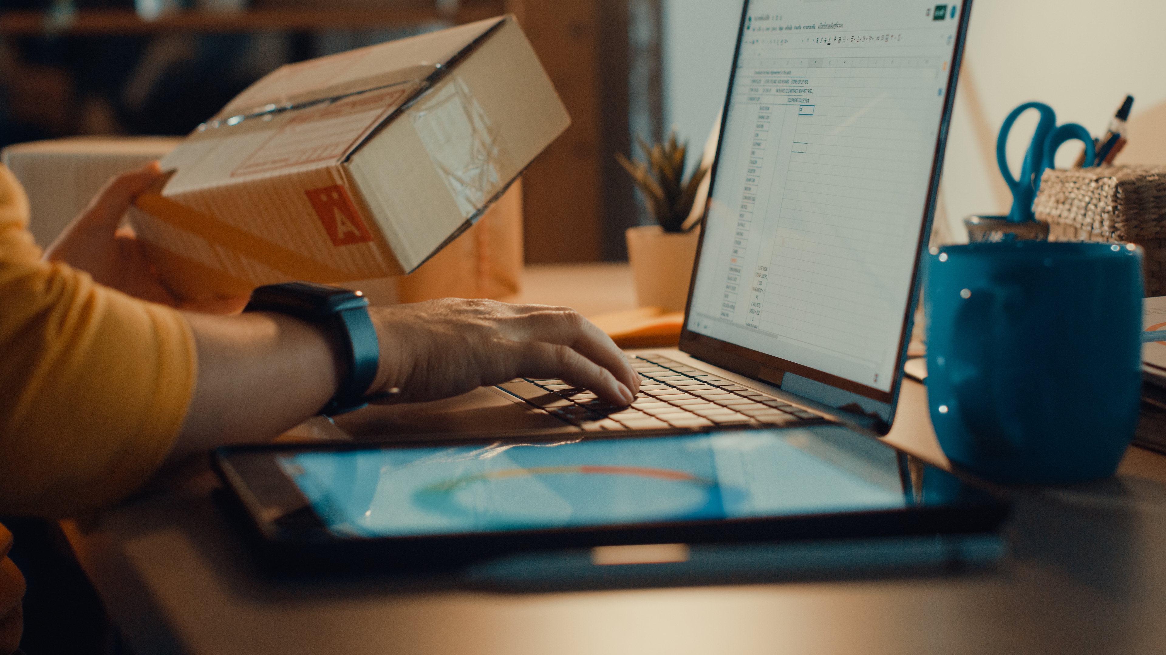 A person working on a laptop at a desk while holding a shipping box, indicating a logistics or e-commerce scenario. The laptop screen displays a spreadsheet, and nearby, a tablet, coffee mug, and various office supplies are visible, suggesting a multitasking work environment.