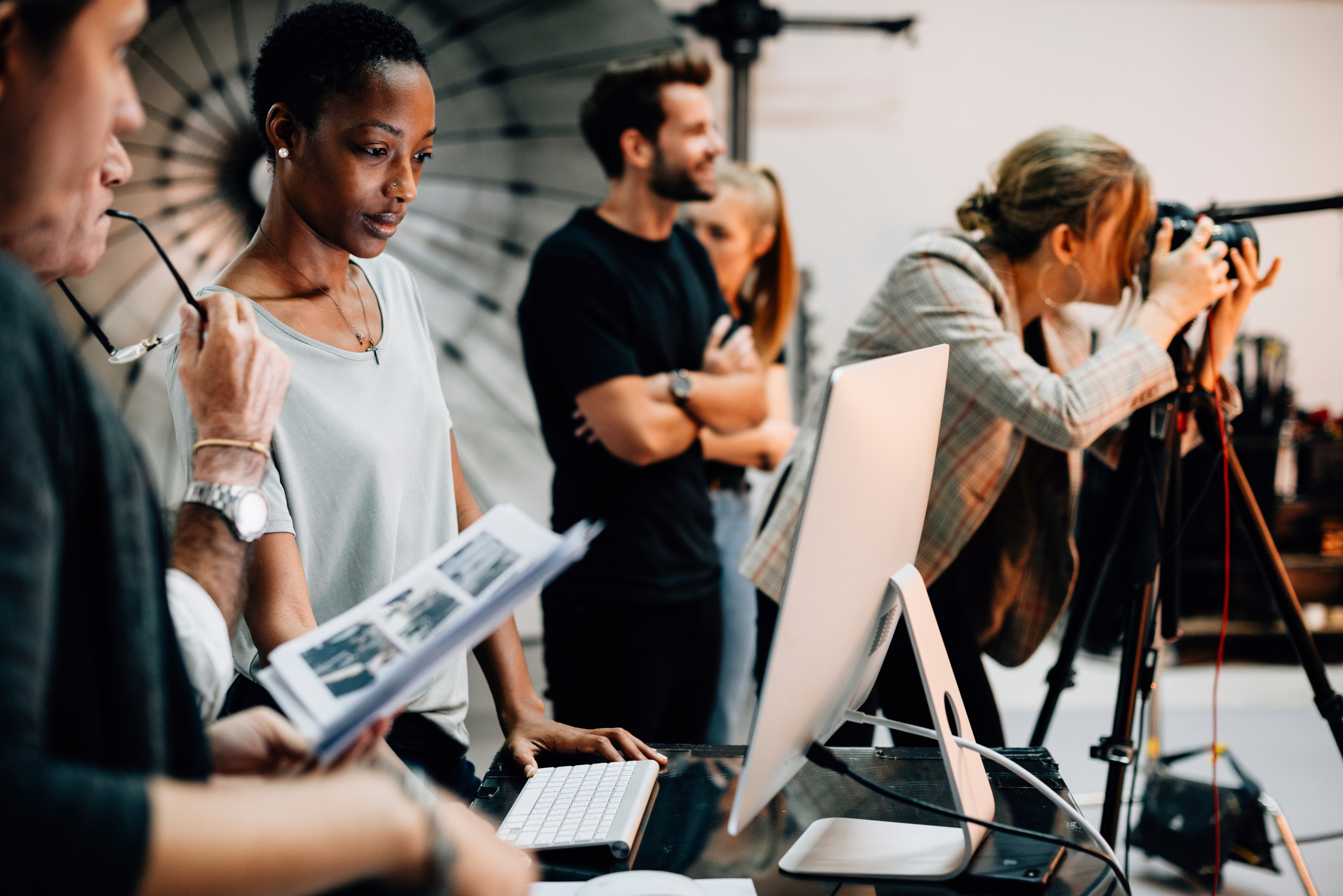 A group of creative professionals working in a photography studio. One person is reviewing images on a computer, while another photographer is actively taking photos using a camera on a tripod. The team is engaged in a collaborative process, with lighting equipment visible in the background.