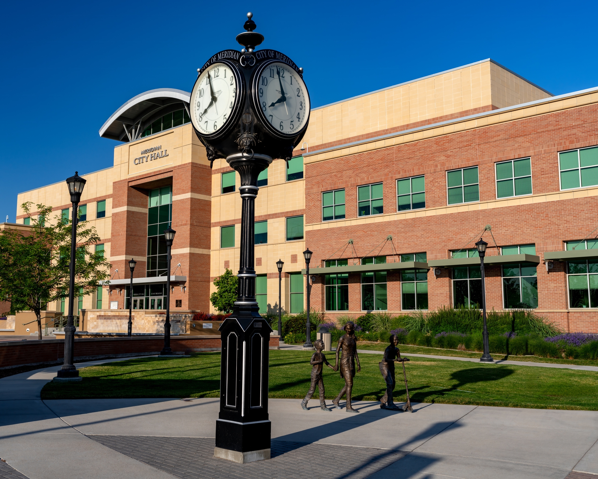 The Meridian City Hall building, framed by a large black clock and bronze sculptures of a walking family, surrounded by a well-maintained lawn and lampposts on a sunny day.