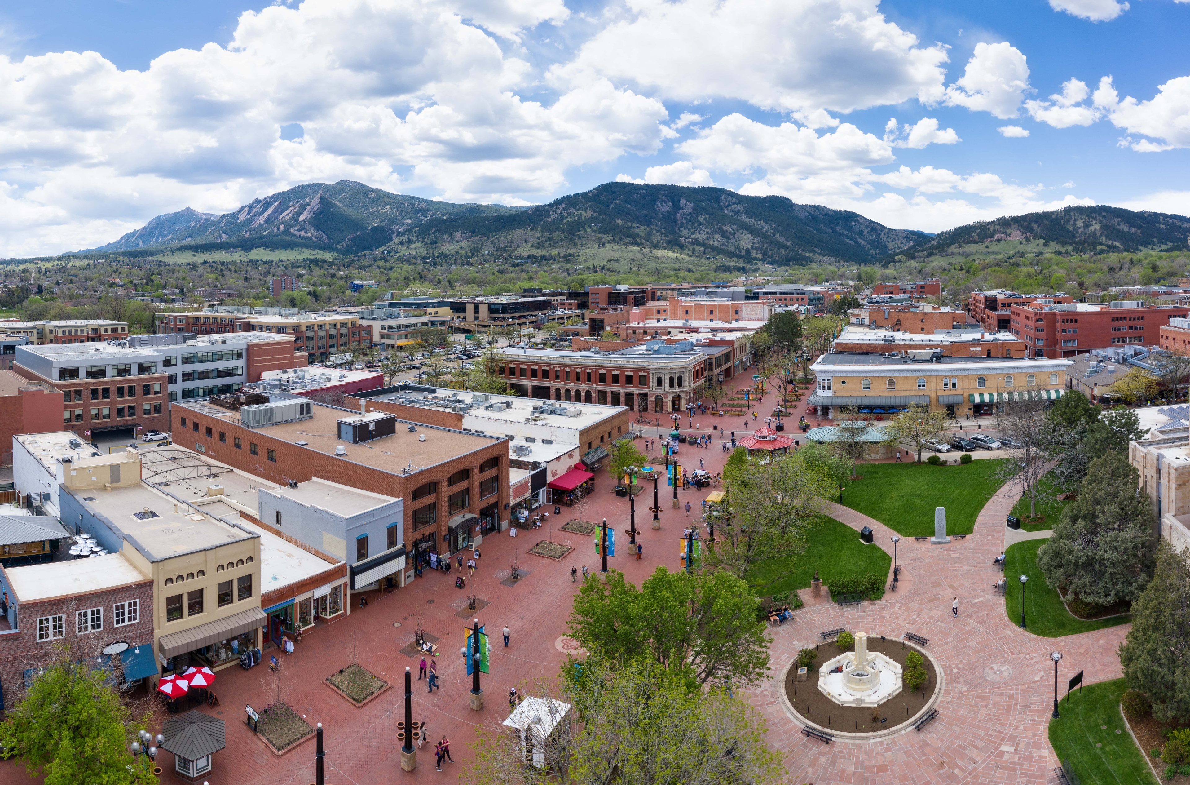Aerial view of downtown Boulder, Colorado, featuring red-brick pedestrian streets, historic buildings, a central plaza with a fountain, and scenic mountains in the background under a partly cloudy sky.