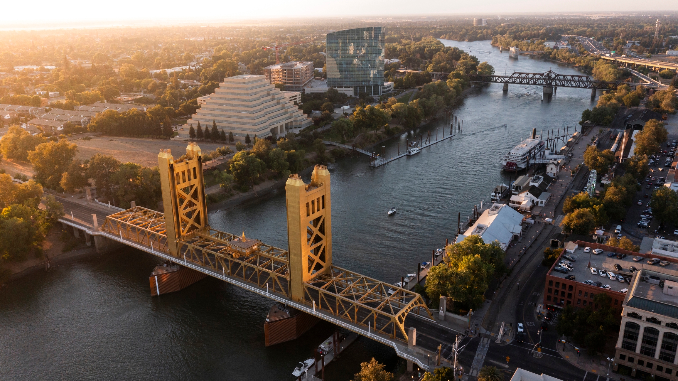 An aerial view of the Tower Bridge in Sacramento, California. The yellow bridge spans the Sacramento River, with a backdrop of modern buildings, including a pyramid-shaped structure, and lush greenery. Boats are visible on the river, and a riverside walkway with small structures and parking areas is on the right.