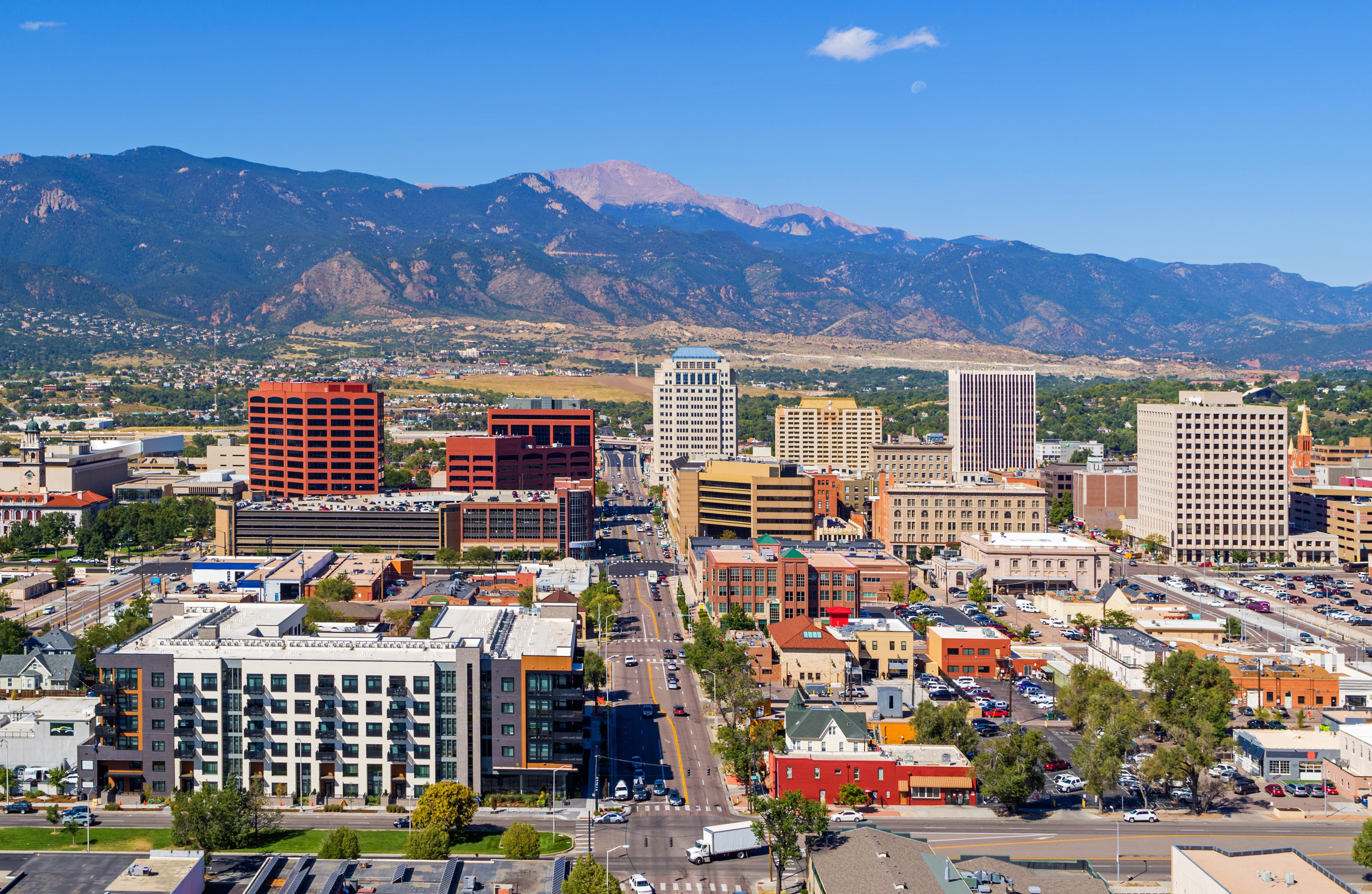 Aerial view of downtown Colorado Springs with Pikes Peak rising in the background, featuring a mix of modern and historic buildings under a clear blue sky.