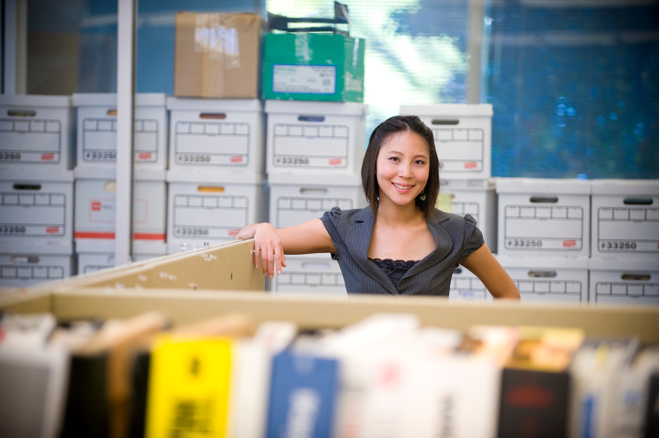 A professional woman smiling confidently in an office setting, surrounded by organized storage boxes in the background, with a partition and blurred books in the foreground.