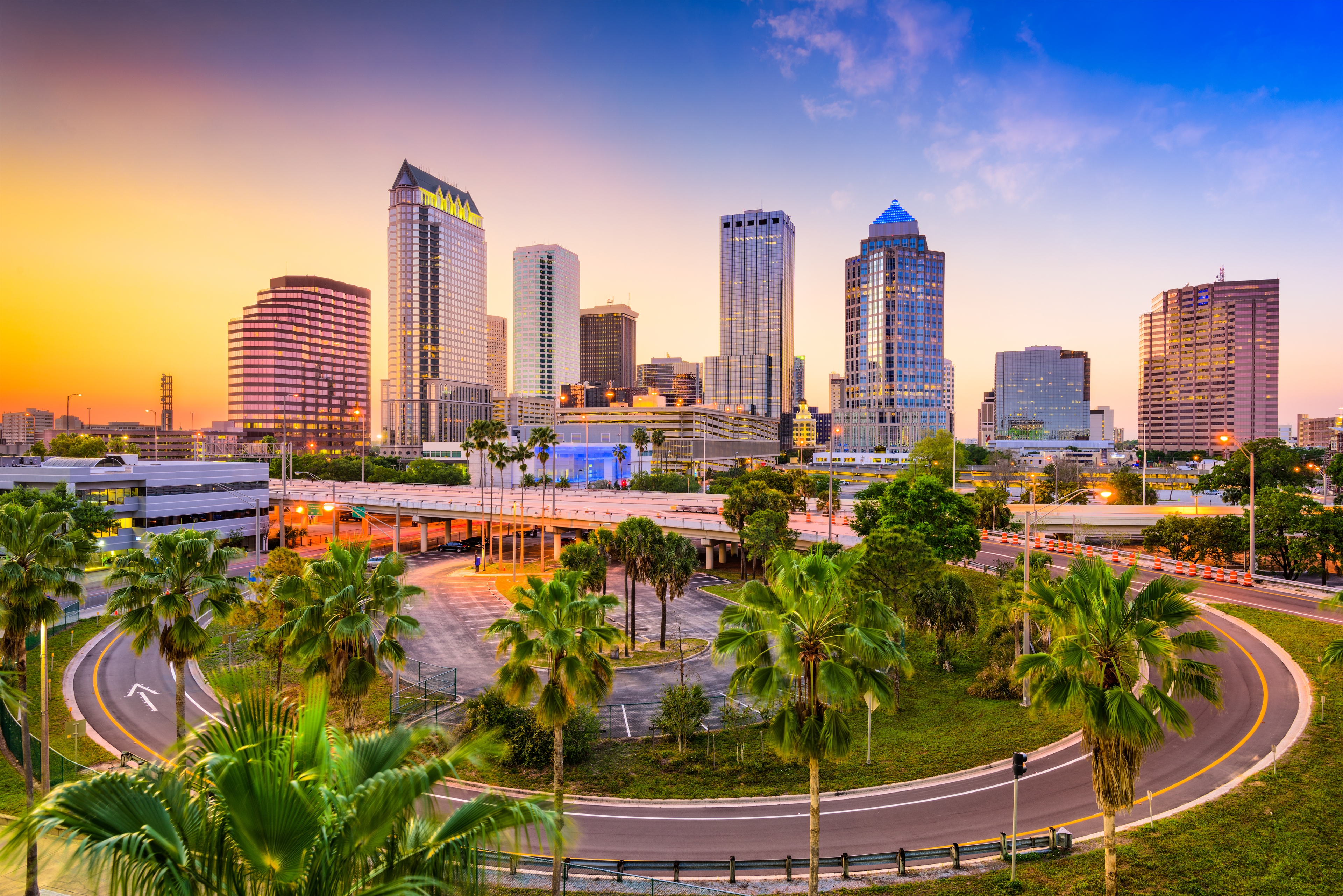 Downtown Tampa skyline at sunset with modern high-rises, palm trees, and a curving roadway in the foreground.