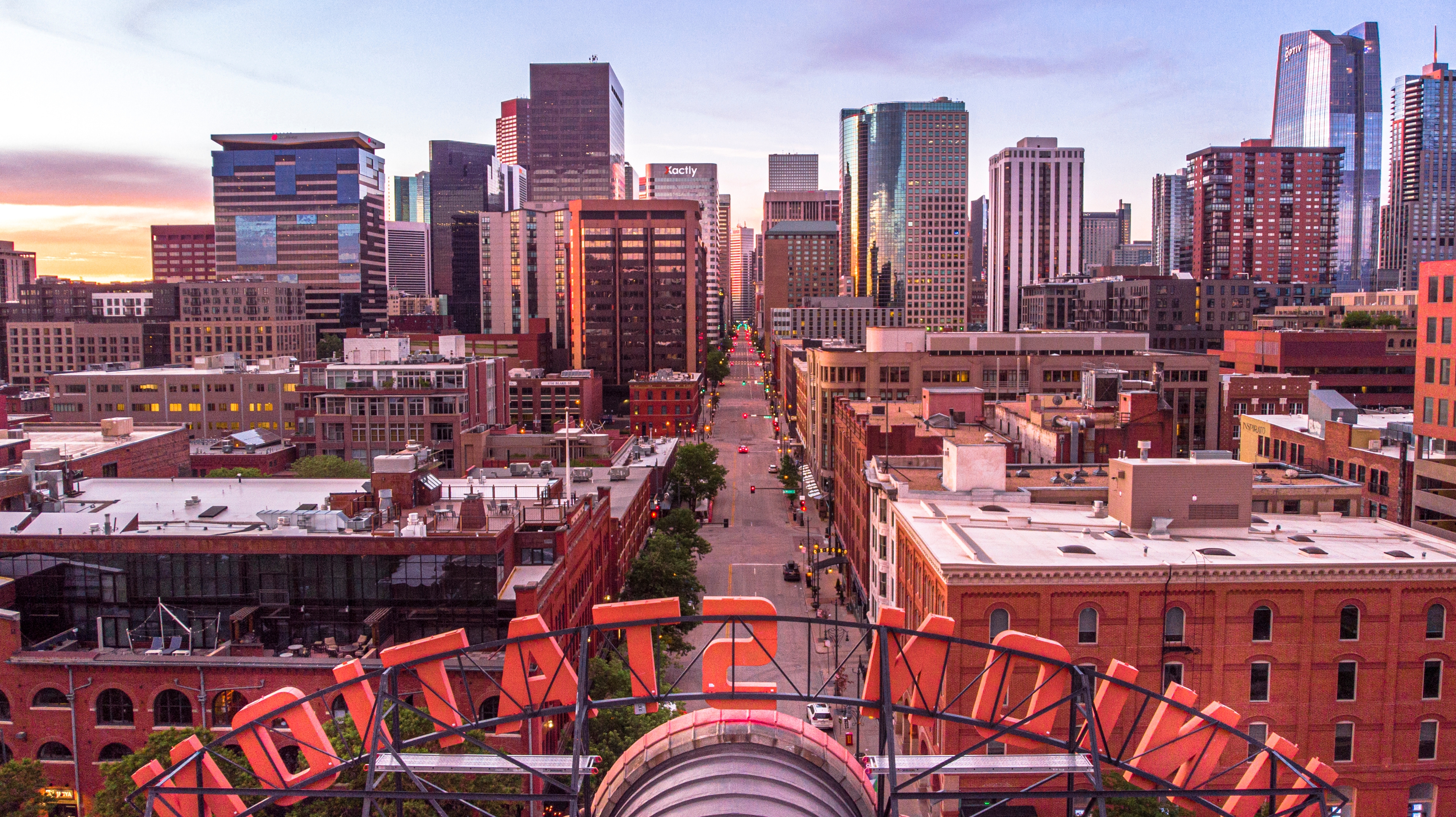 Downtown Denver at dusk with vibrant city lights reflecting off high-rises, historic brick buildings in the foreground, and a neon sign framing the urban skyline.