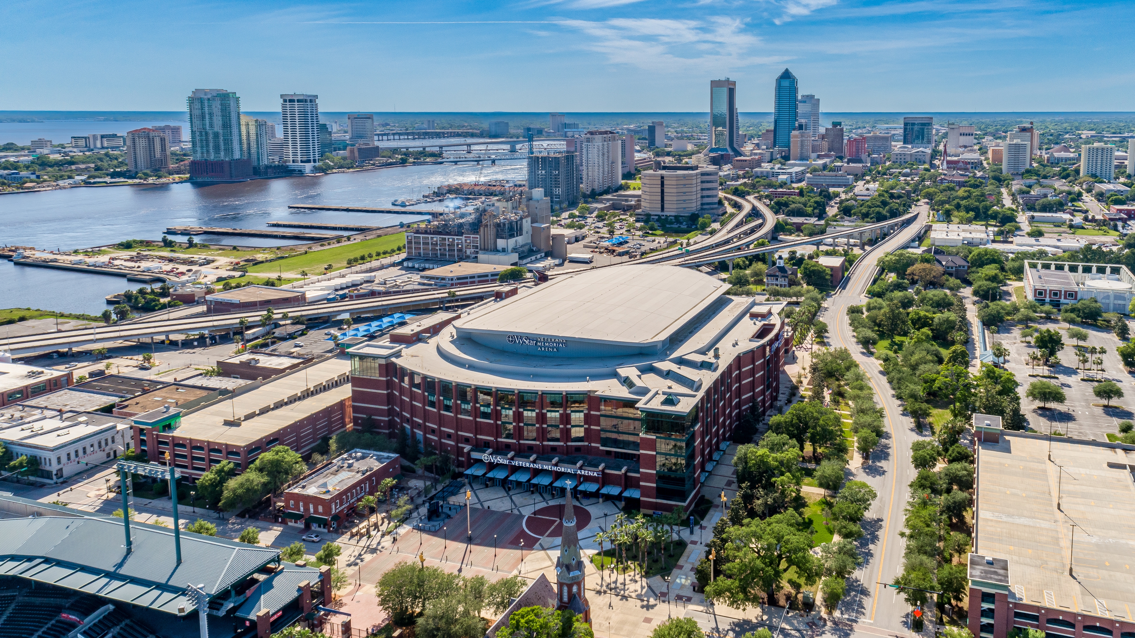 Aerial view of downtown Jacksonville, Florida, showcasing the VyStar Veterans Memorial Arena, the St. Johns River, and a modern skyline under a clear blue sky.