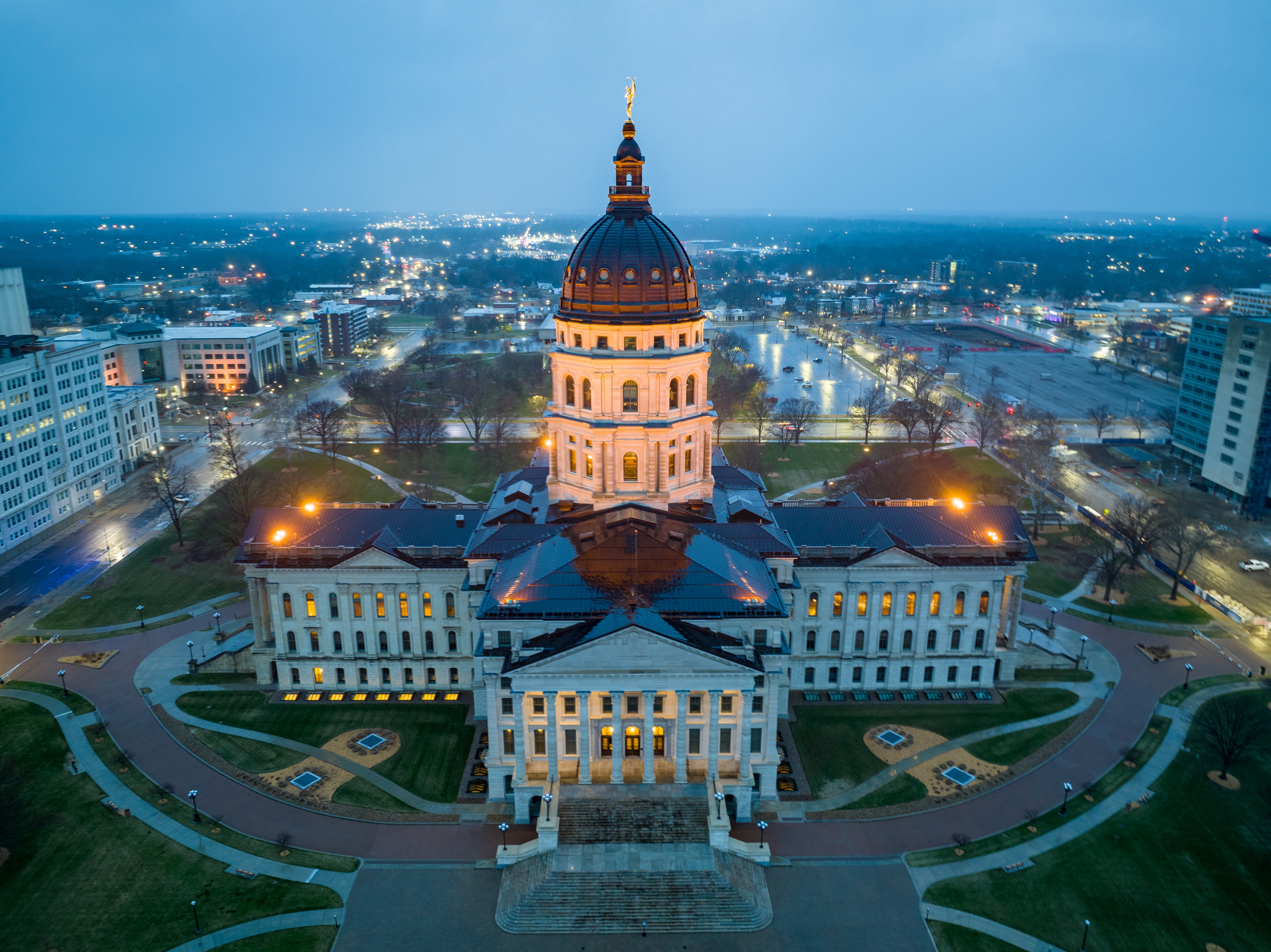 Aerial view of the Kansas State Capitol building illuminated at dusk in Topeka, Kansas, with the surrounding downtown cityscape.
