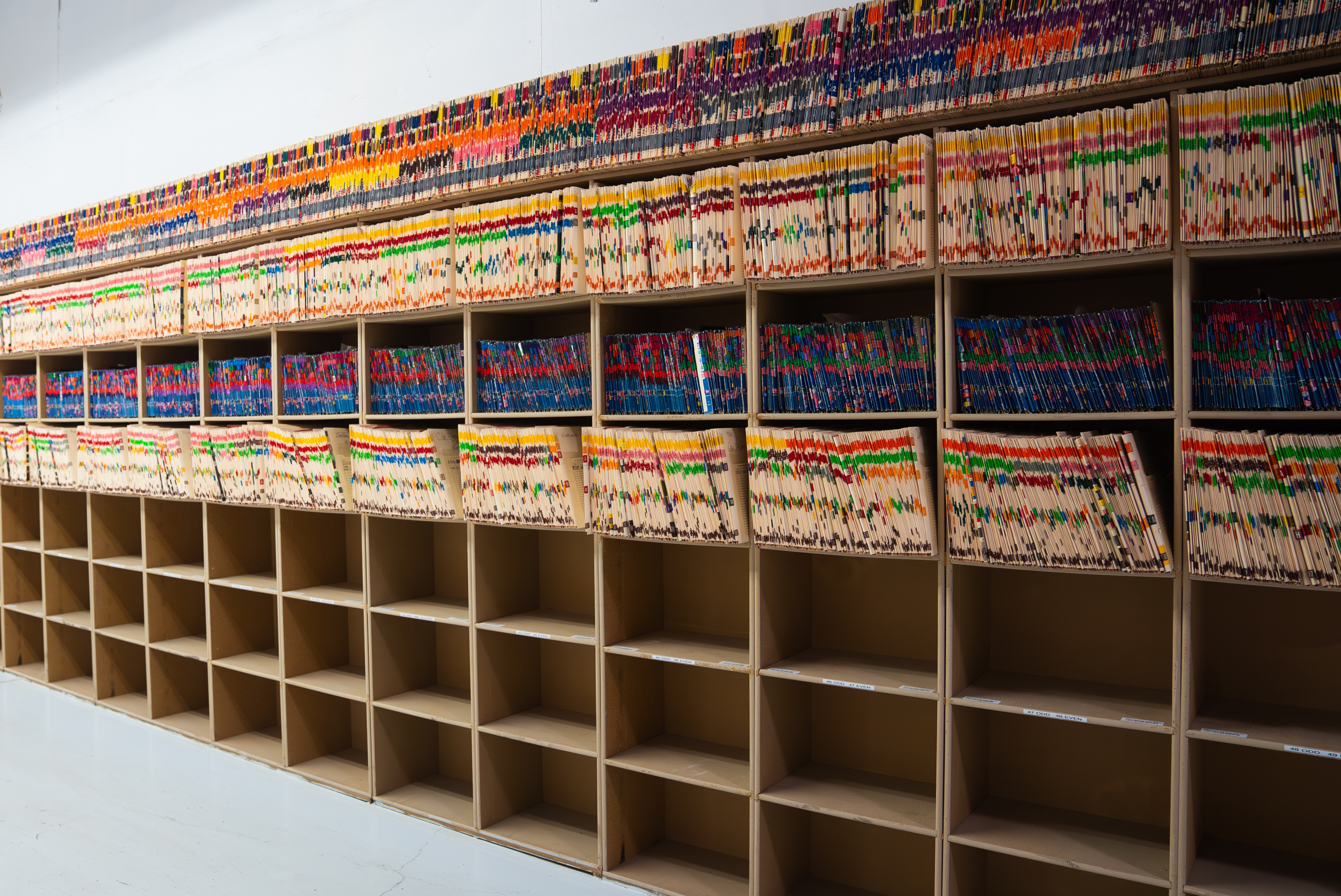 Wall of organized, colorful file folders on shelves in a records room, with several empty slots below