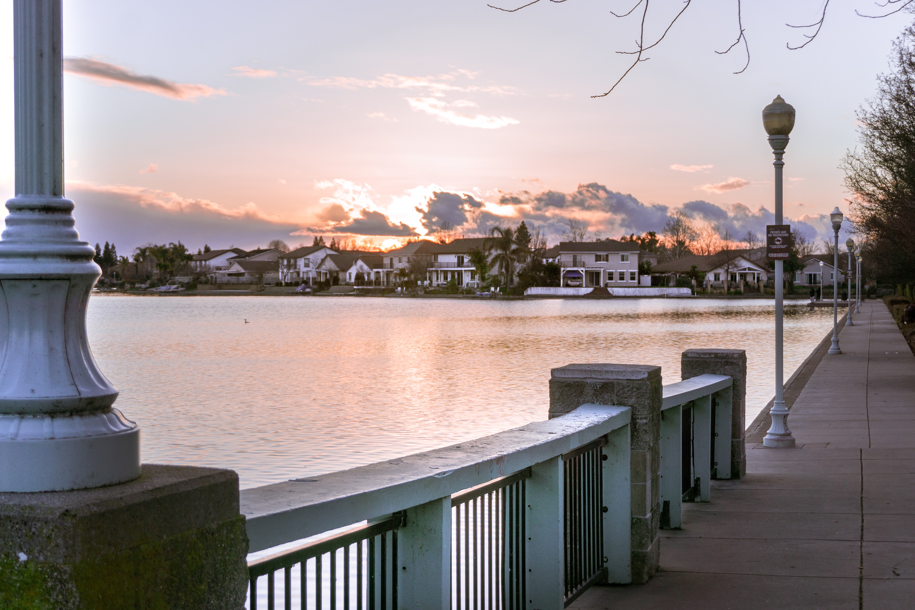 A lakeside walkway in Elk Grove, California, featuring street lamps and a railing along the water, with houses reflecting on the lake under a colorful sunset sky.