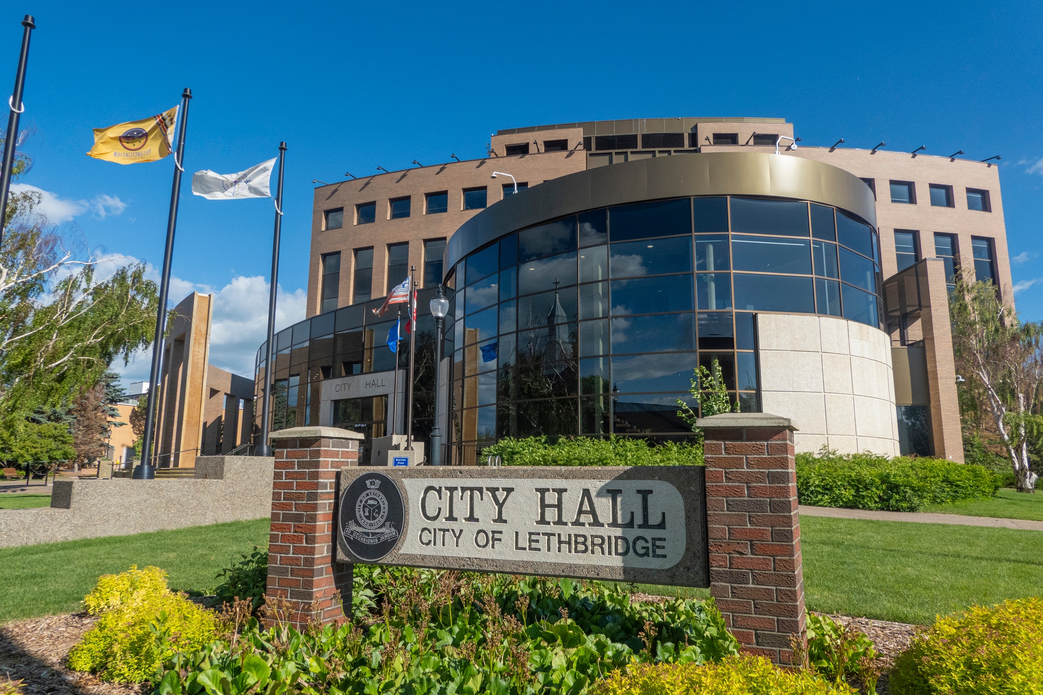 A sunny view of Lethbridge City Hall in Alberta, Canada, featuring its modern glass and brick facade, a landscaped foreground, and flags waving against a bright blue sky.