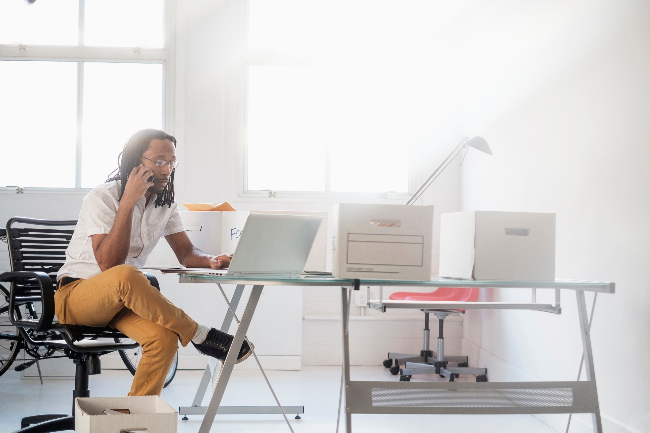 A professional working in a minimalist office environment, seated at a glass desk with a laptop and phone. The space is bright, with natural light streaming through large windows, and is organized with storage boxes for files and documents.