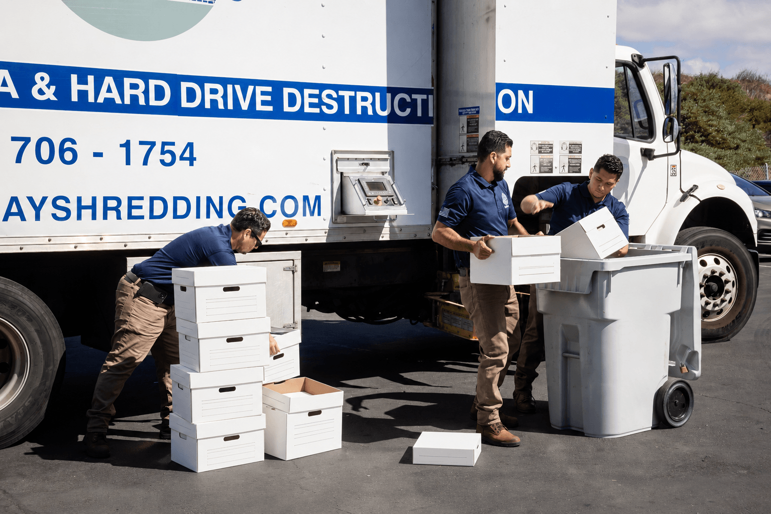 Mobile shredding service team loading document boxes into a secure shredding bin next to a hard drive destruction truck during an on-site shredding event.