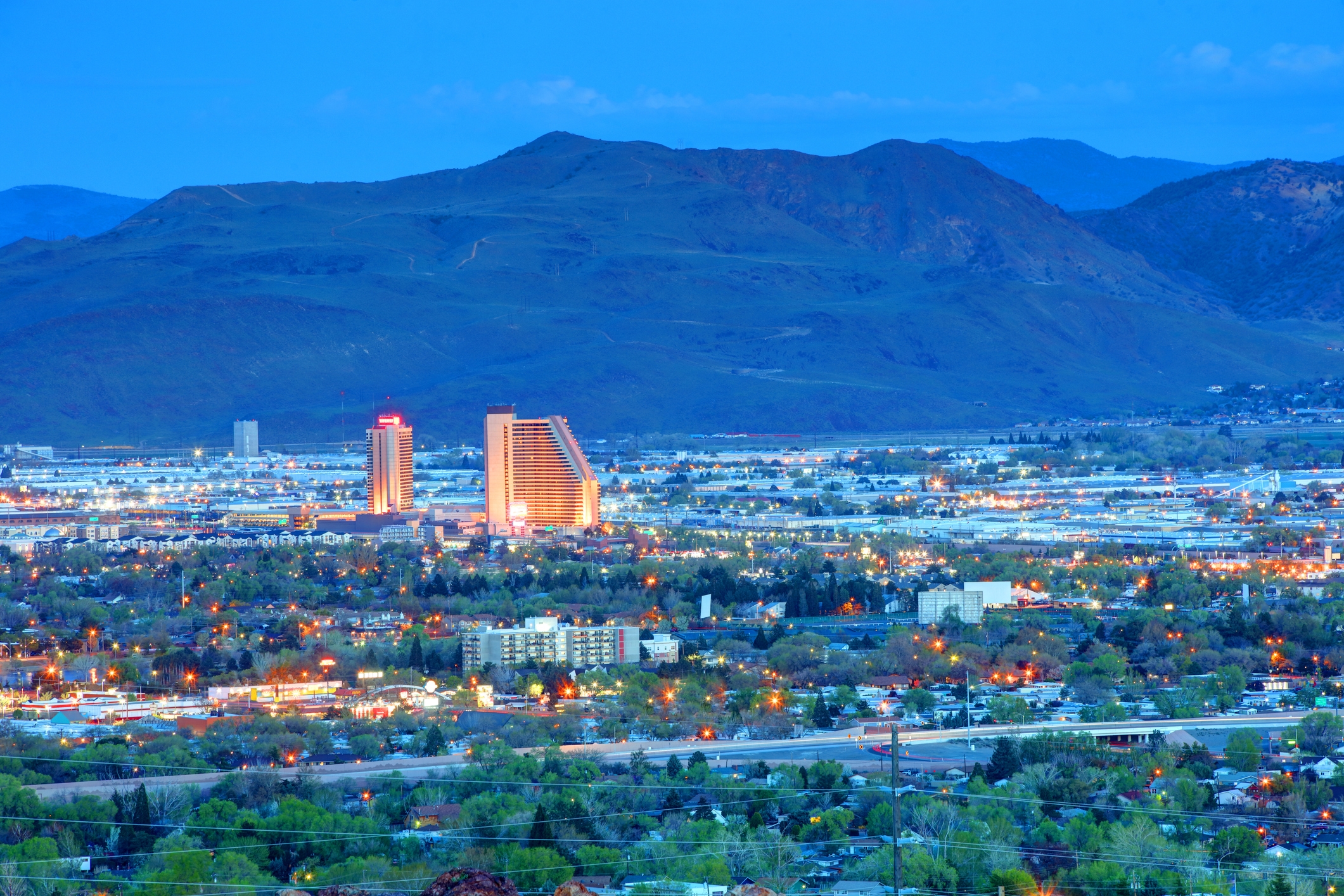 An evening view of Sparks, Nevada, with illuminated buildings and streets spread across the valley. Two brightly lit hotel towers stand out in the foreground.