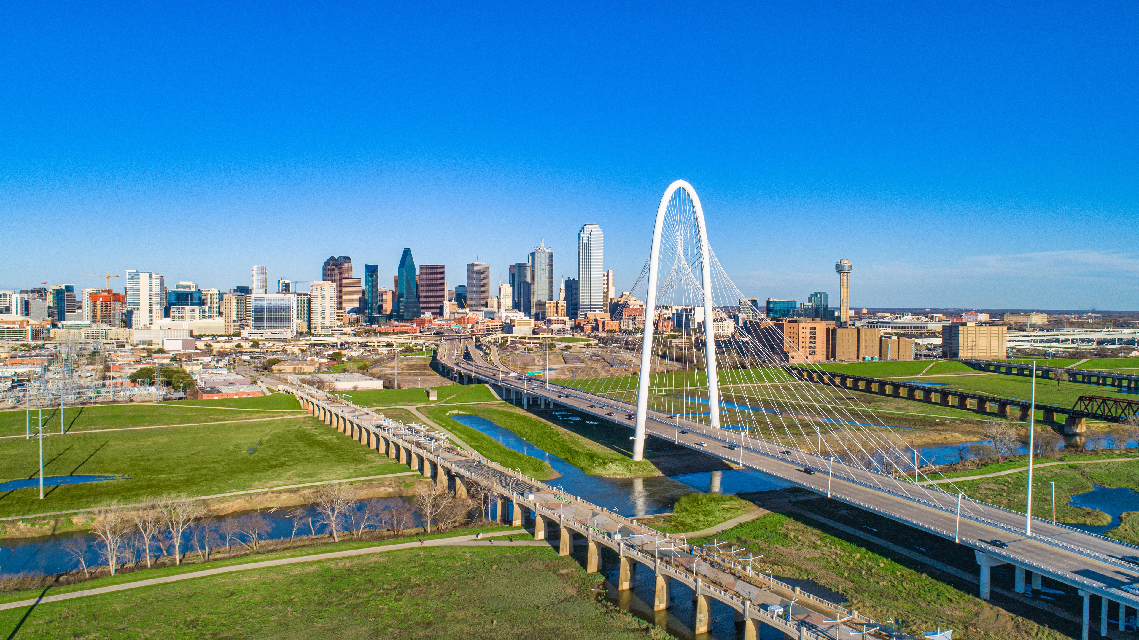 Panoramic view of Dallas, Texas, featuring the iconic Margaret Hunt Hill Bridge in the foreground and the city’s modern skyline under a clear blue sky.