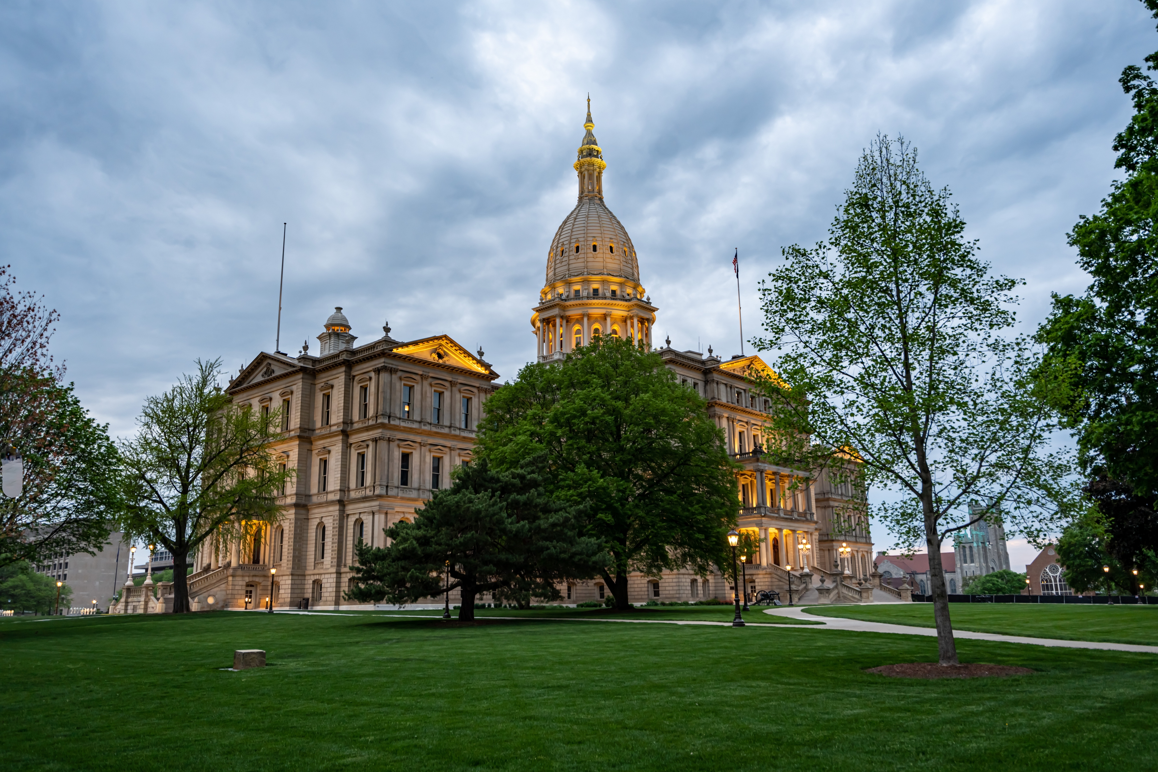 The Michigan State Capitol in Lansing illuminated at dusk, showcasing its distinctive dome and neoclassical architecture, surrounded by green lawns and trees under a cloudy sky.