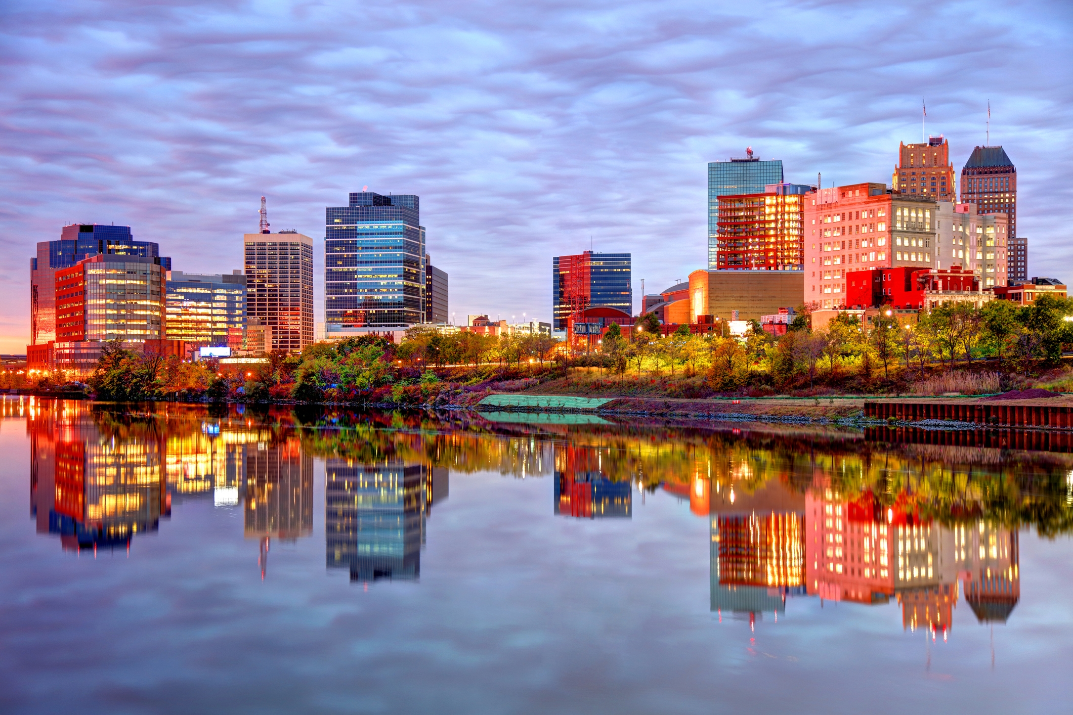 An evening view of Newark, New Jersey, featuring a mix of contemporary high-rise buildings and historic architecture along the riverfront. Brightly lit windows and colorful lighting reflect vividly on the still water, with a row of trees and green spaces lining the shoreline under a textured, cloud-filled sky.