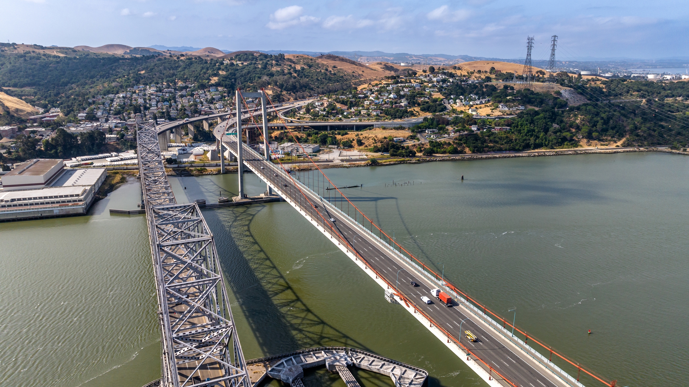 An aerial view of the Carquinez Bridge in Vallejo, California, spanning across the Carquinez Strait. The bridge features two distinct structures, including a suspension design and a cantilever style. Surrounding hills with residential homes and scattered greenery provide a scenic backdrop. 