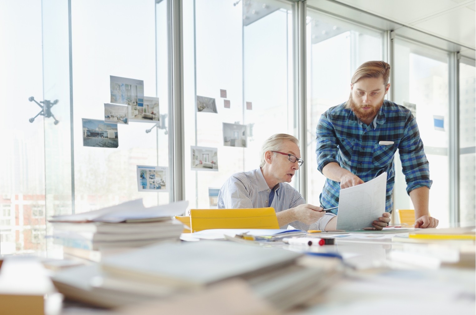 Two real estate managers reviewing blueprints in a bright, modern office with large windows, surrounded by design materials and photos pinned to glass walls.