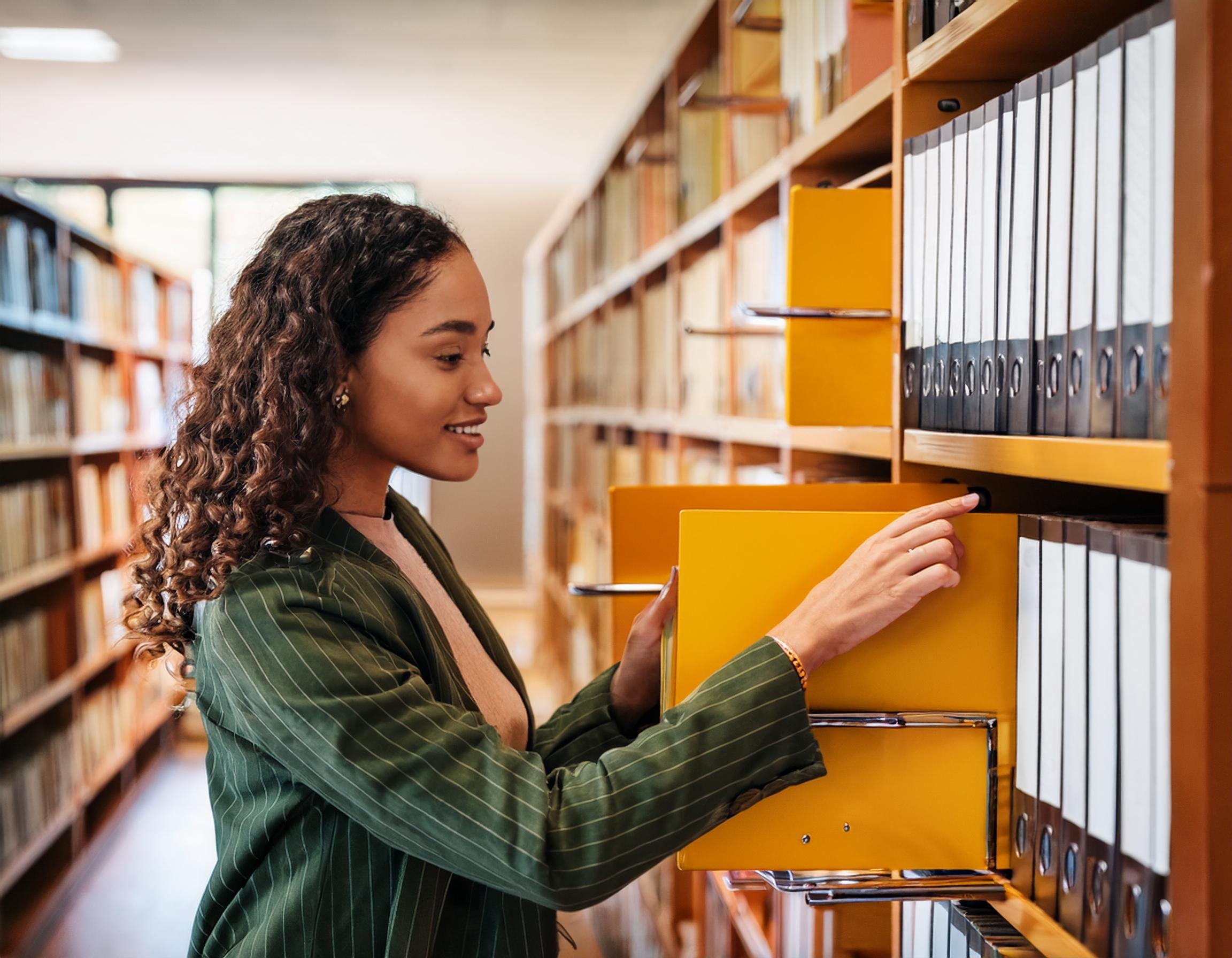 A young professional woman in a green striped blazer browsing through yellow file folders in a library or archive setting, surrounded by shelves filled with organized binders.