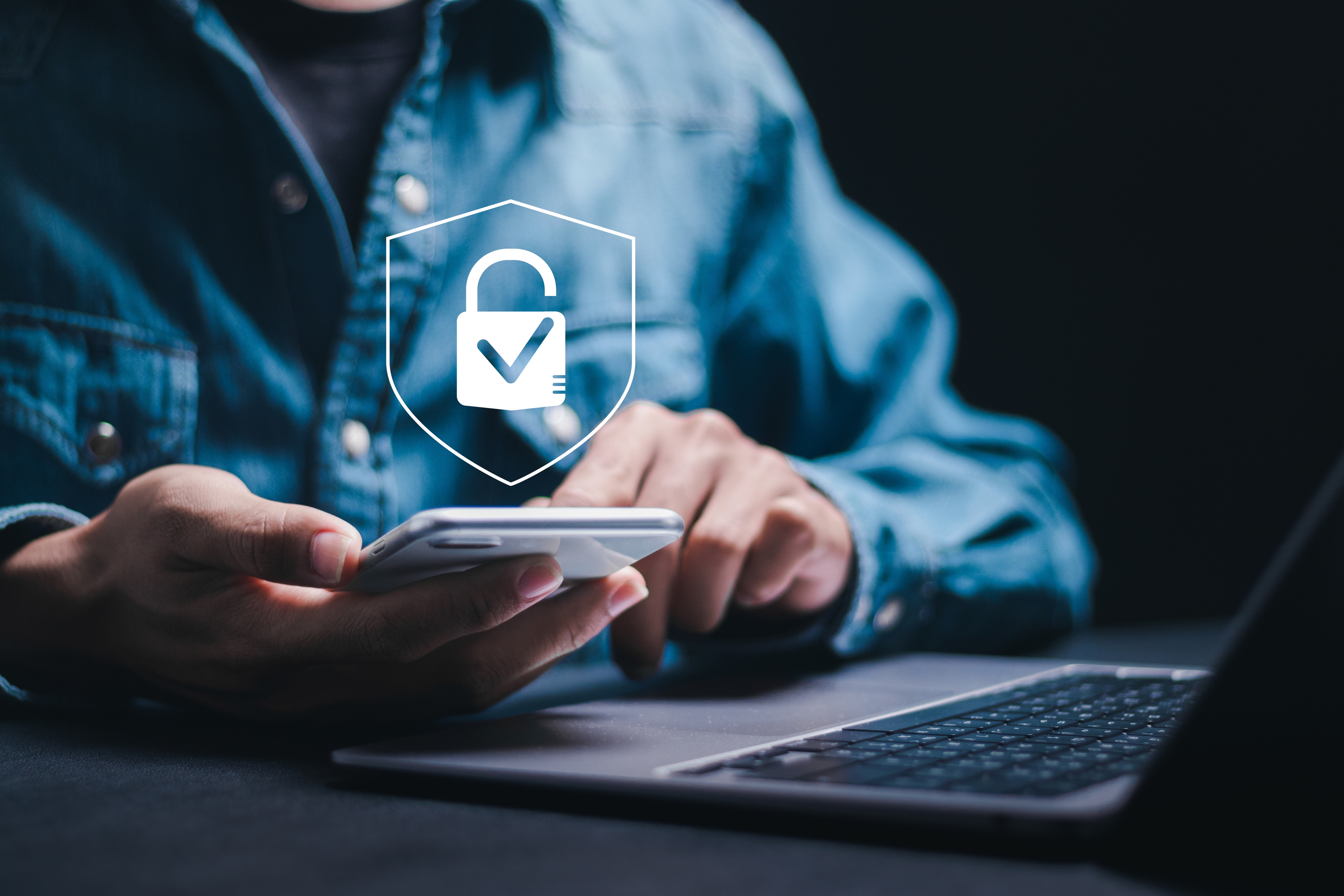 A person using a smartphone with a security icon overlay, depicting a lock with a check mark, symbolizing secure communication or data protection. The individual is sitting at a desk, with a laptop partially visible next to the phone.