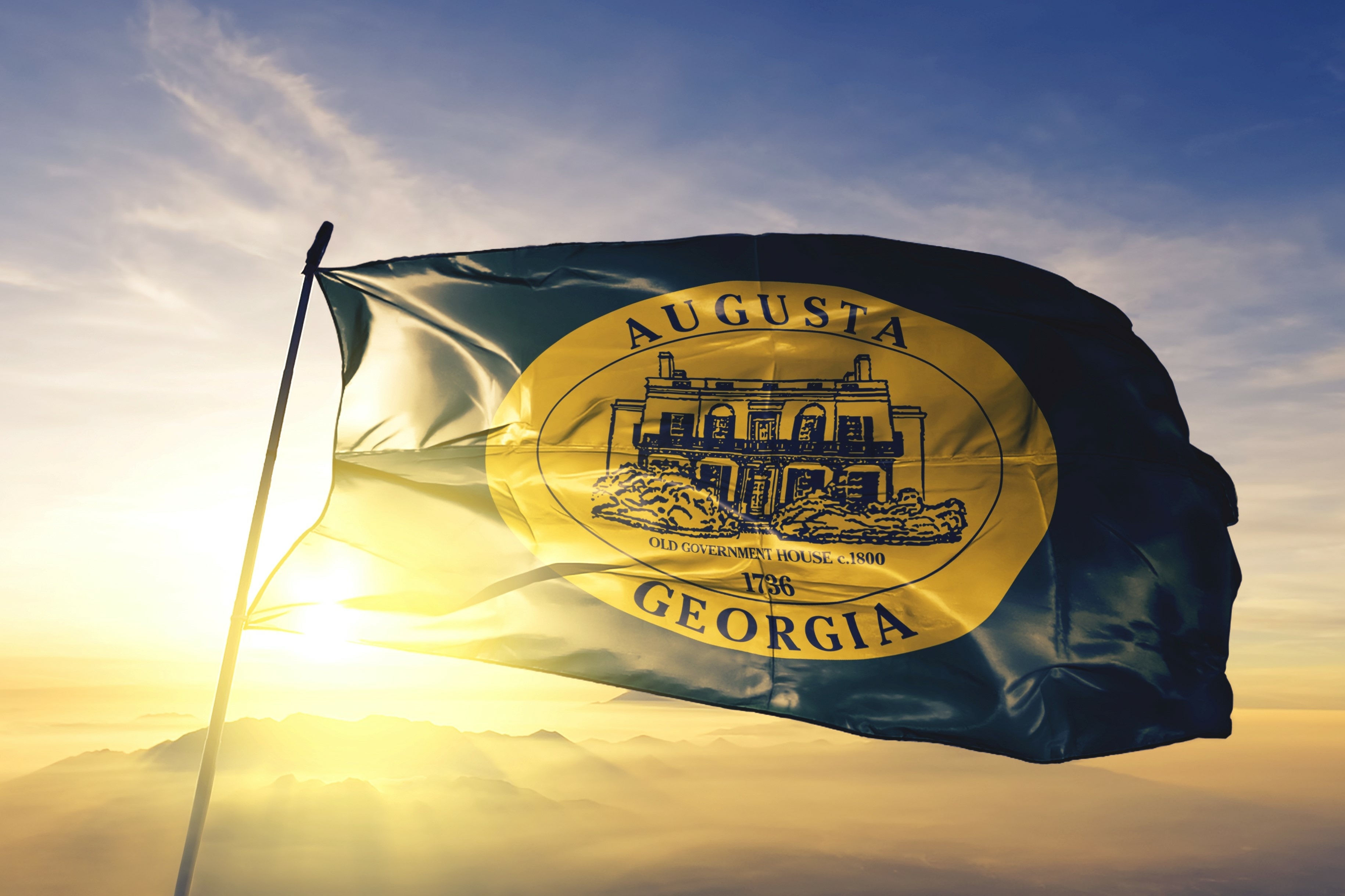 Flag of Augusta, Georgia waving at sunrise, featuring the Old Government House emblem against a backdrop of golden light and mountain silhouettes.