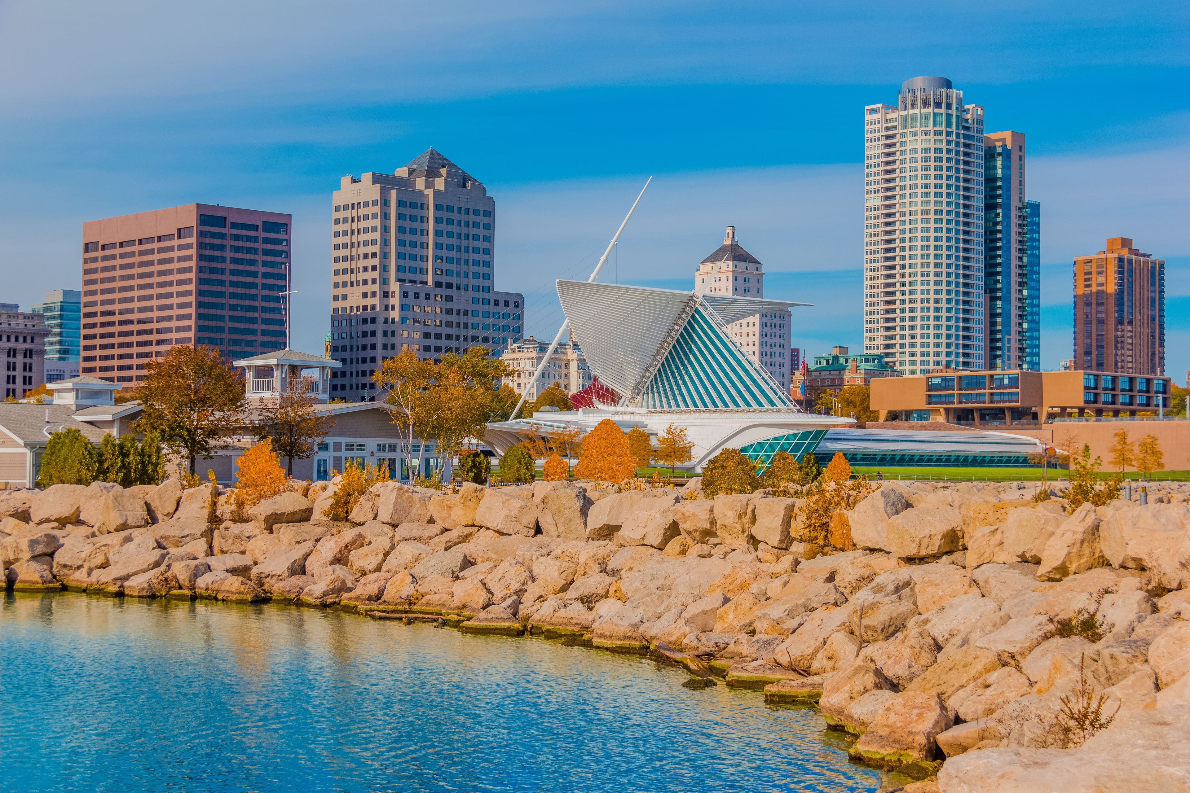 Daytime view of the Milwaukee skyline with the Milwaukee Art Museum's winged architecture in the foreground, surrounded by fall foliage and Lake Michigan’s rocky shoreline.
