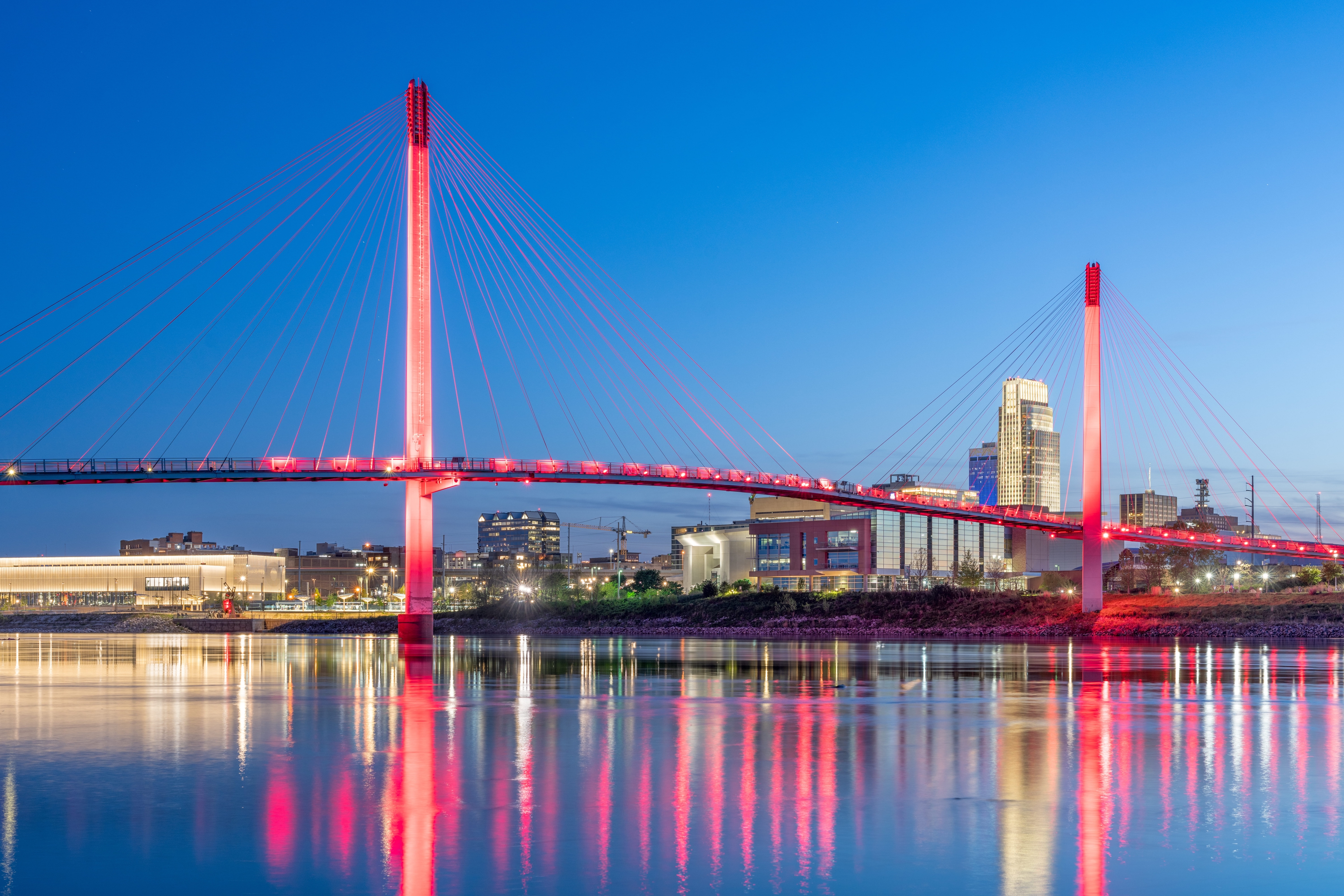Illuminated pedestrian bridge over the Missouri River with the Omaha, Nebraska skyline reflected in the water at dusk.