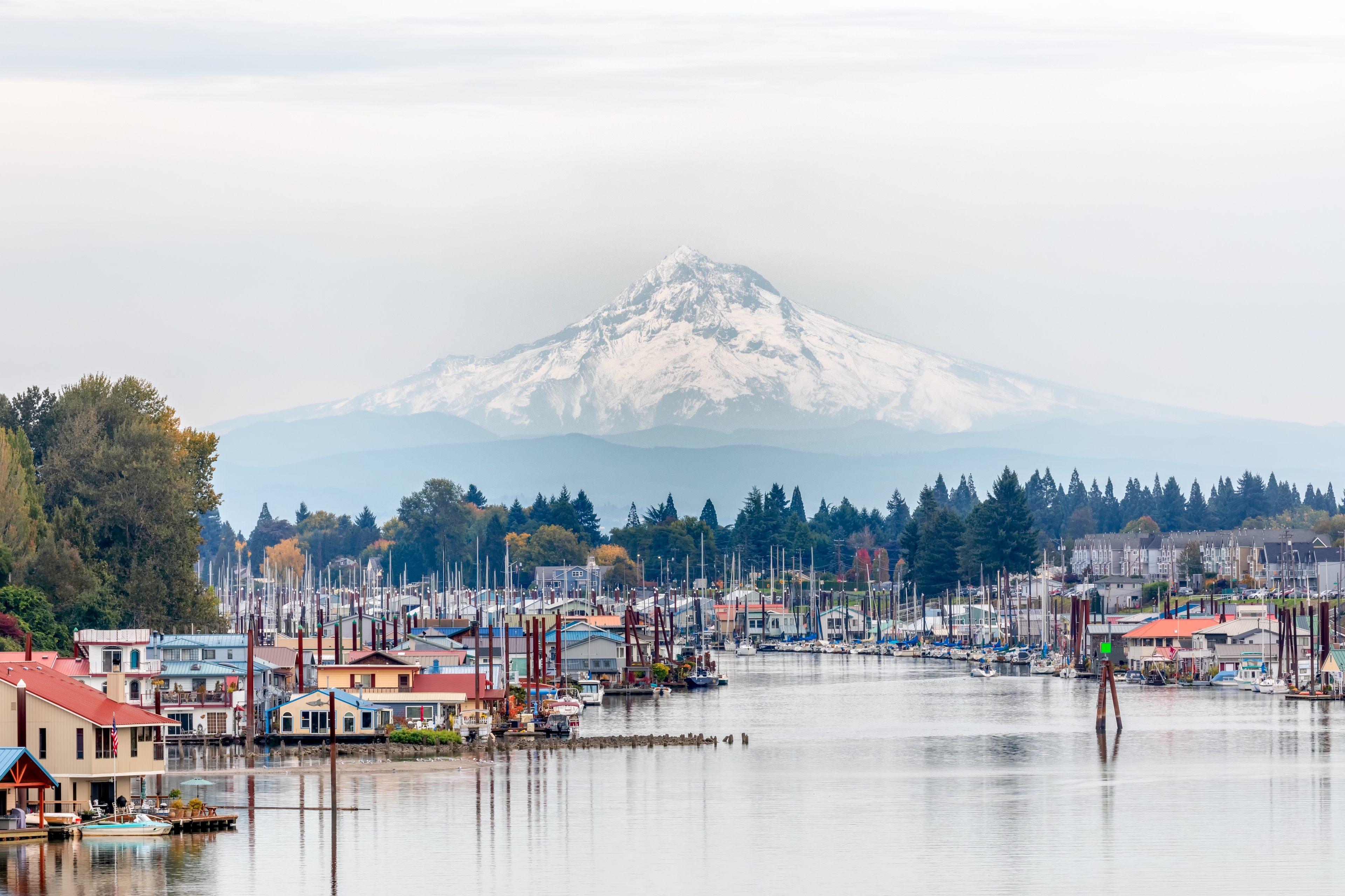 Floating homes and marina on a calm river in Portland, Oregon, with snow-capped Mount Hood rising in the background beneath an overcast sky.