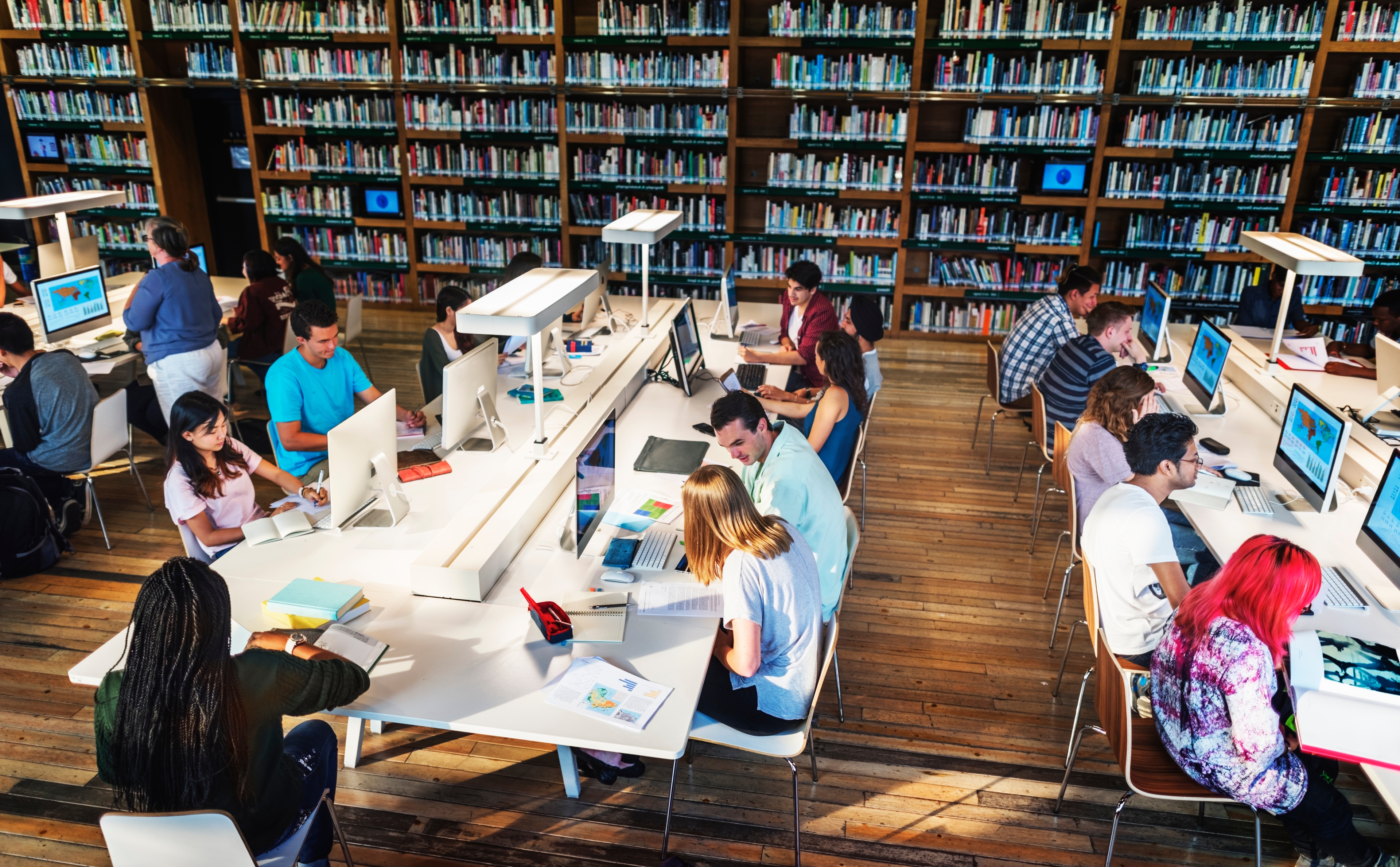 A vibrant library setting with students studying at communal tables, using computers and reading materials, surrounded by bookshelves filled with colorful books.