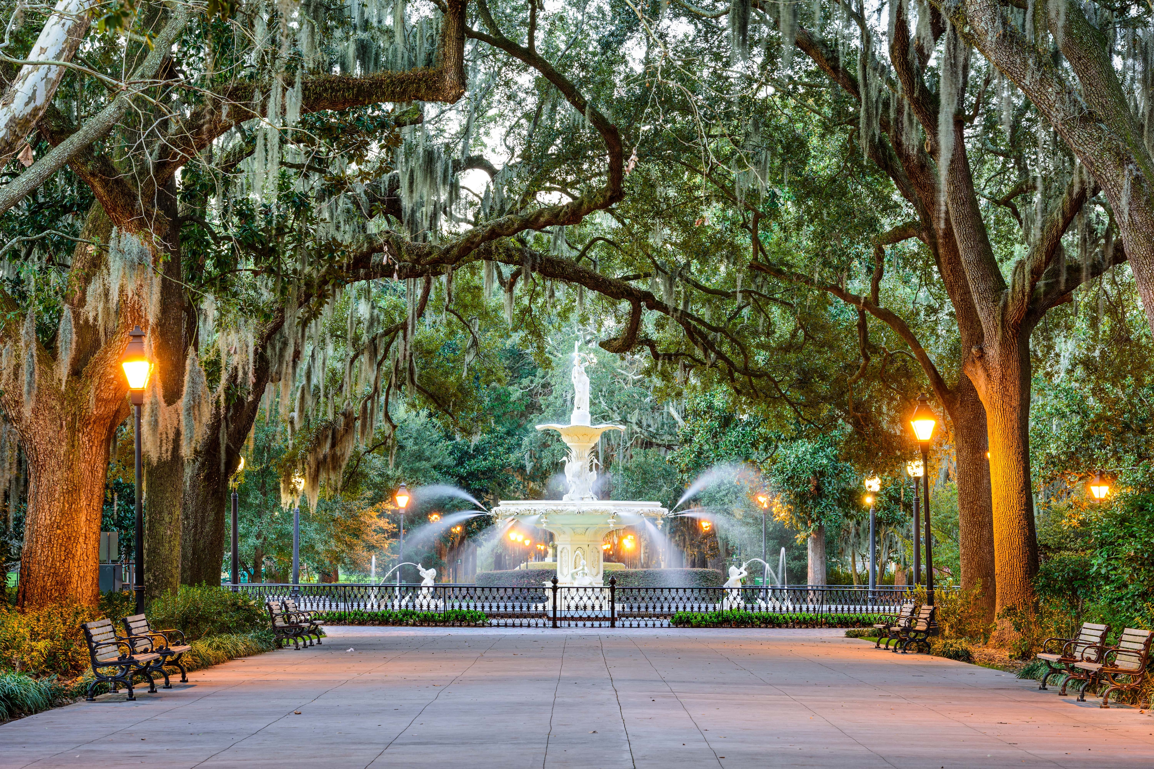 Iconic view of the Forsyth Fountain in Savannah’s Forsyth Park, framed by moss-draped oak trees and glowing lampposts along a quiet, tree-lined promenade.