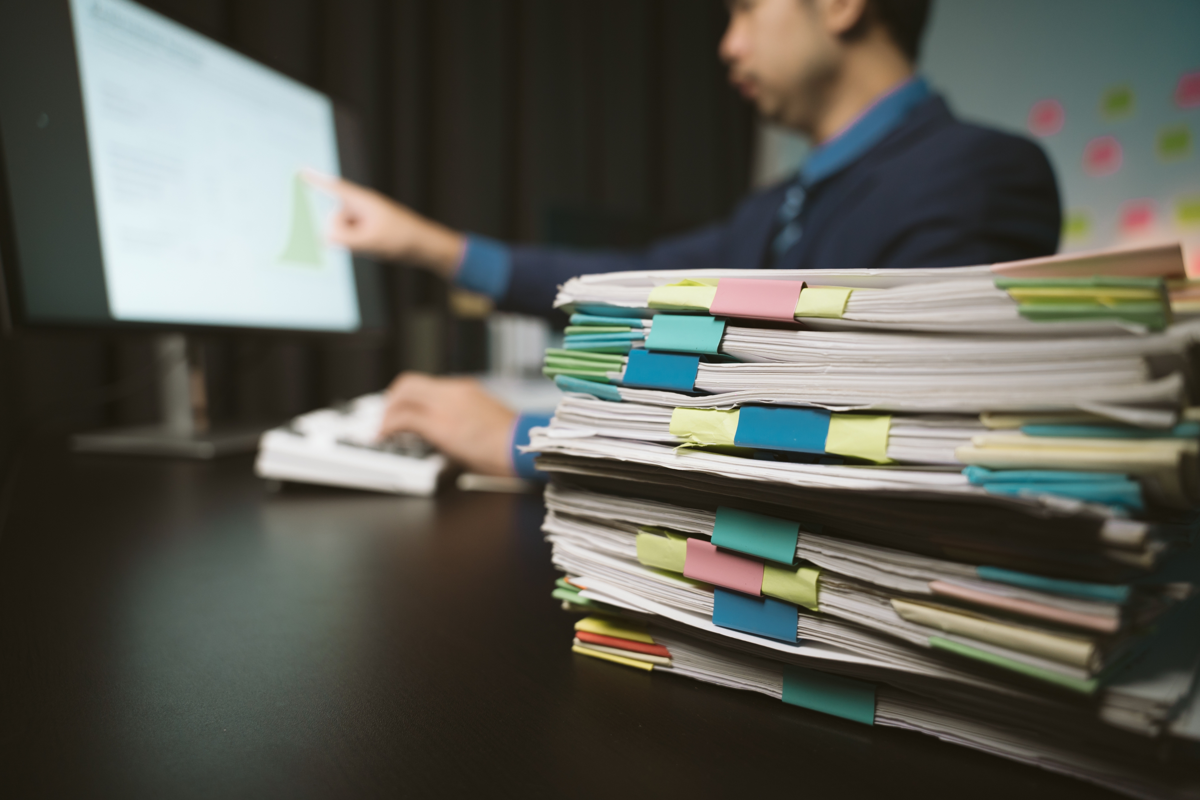 Office worker pointing at a computer screen with a stack of documents organized with colorful clips in the foreground.