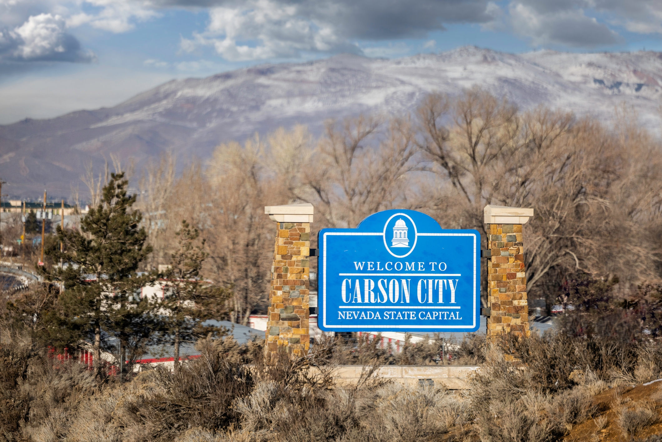 A sign reading 'Welcome to Carson City, Nevada State Capital' stands in front of a landscape with dry brush, bare trees, and distant snow-covered mountains under a partly cloudy sky.