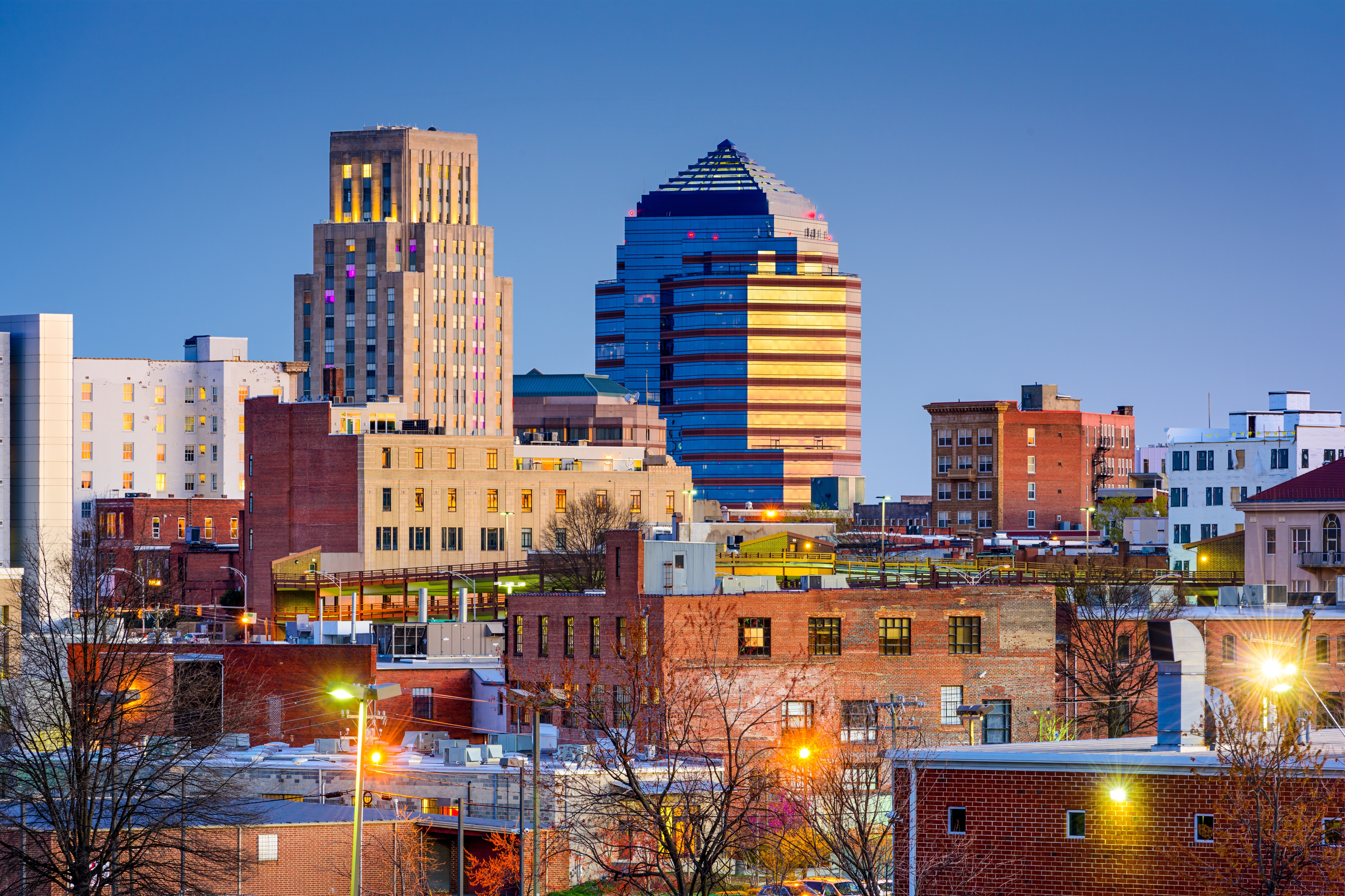 Evening view of downtown Durham, North Carolina, with historic brick buildings in the foreground and the iconic One City Center glowing against a clear twilight sky.