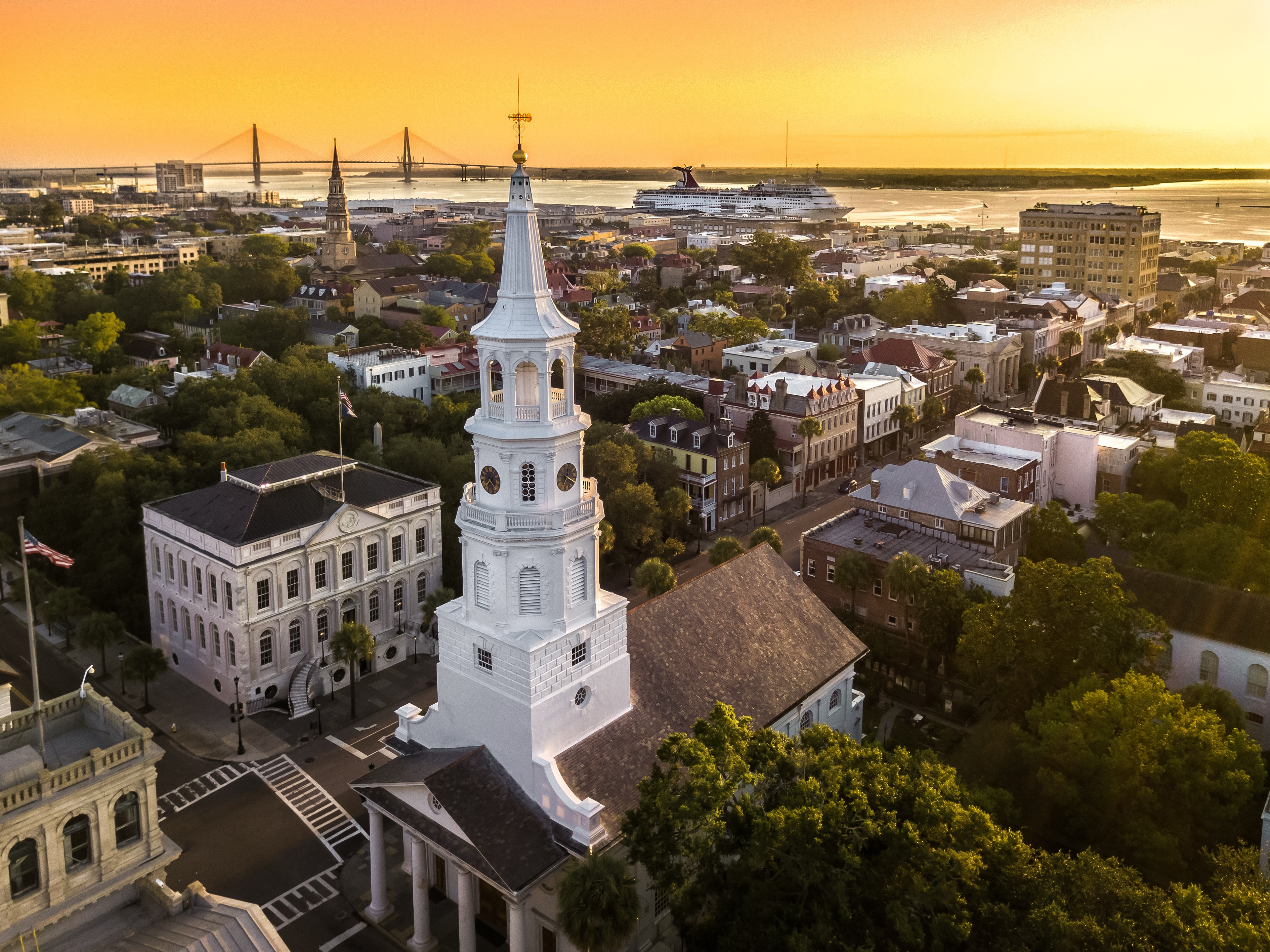 Sunset aerial view of historic downtown Charleston, South Carolina, featuring St. Michael’s Church steeple, colonial architecture, and the Ravenel Bridge in the distance over the harbor.