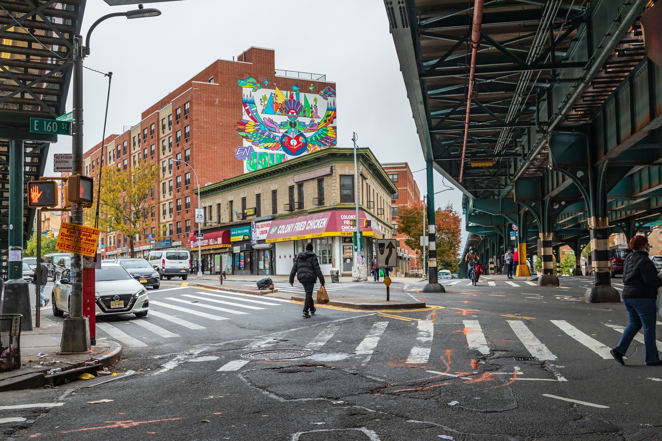 An urban scene in the Bronx, New York, featuring a vibrant mural on a brick apartment building, shops on a street corner, and an elevated subway track. The crosswalk and traffic signal are visible, with people walking and cars passing through the intersection.