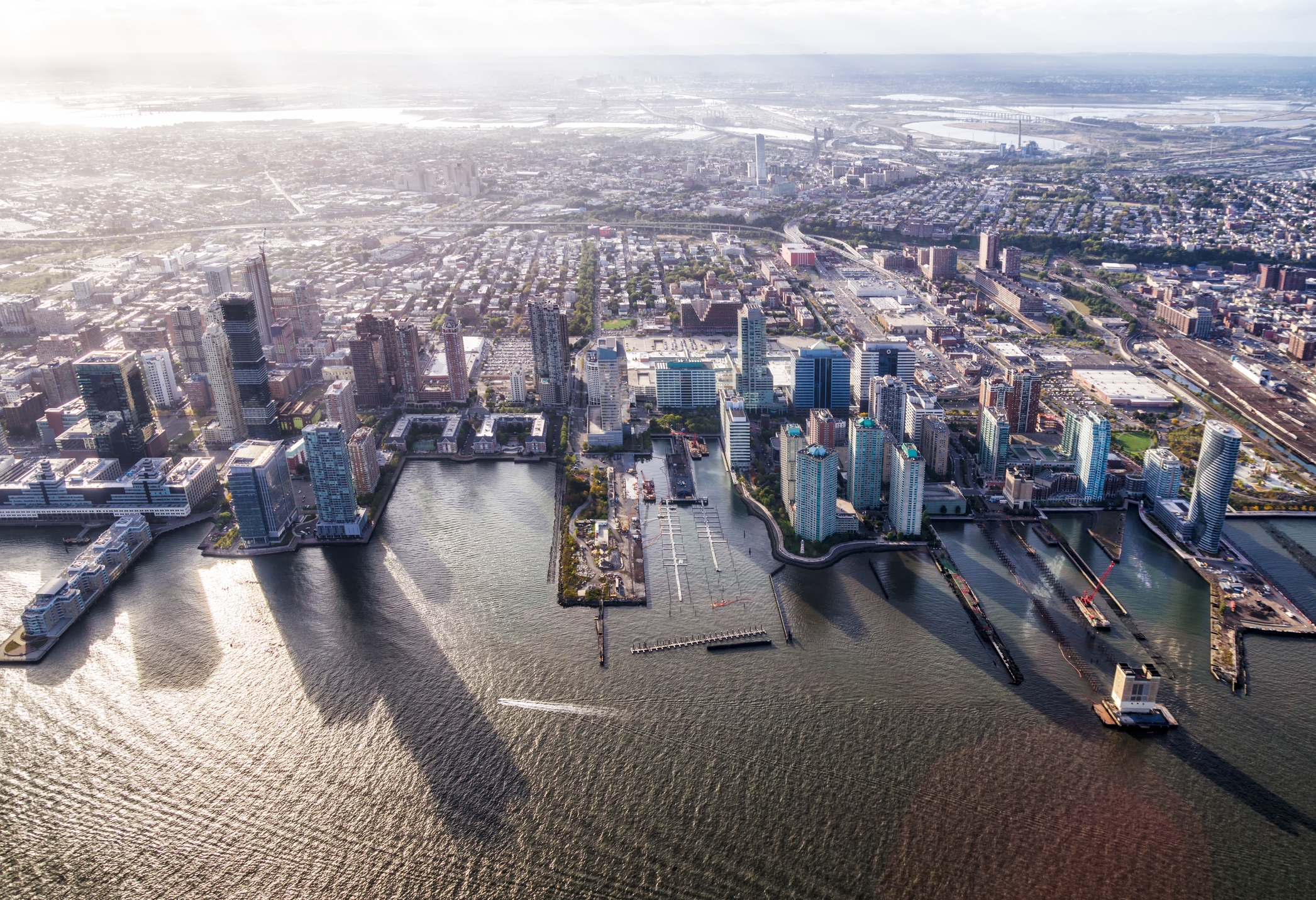 Aerial view of Jersey City, New Jersey, featuring tall buildings by the waterfront, marinas, and piers. The Hudson River and city neighborhoods extend into the distance.