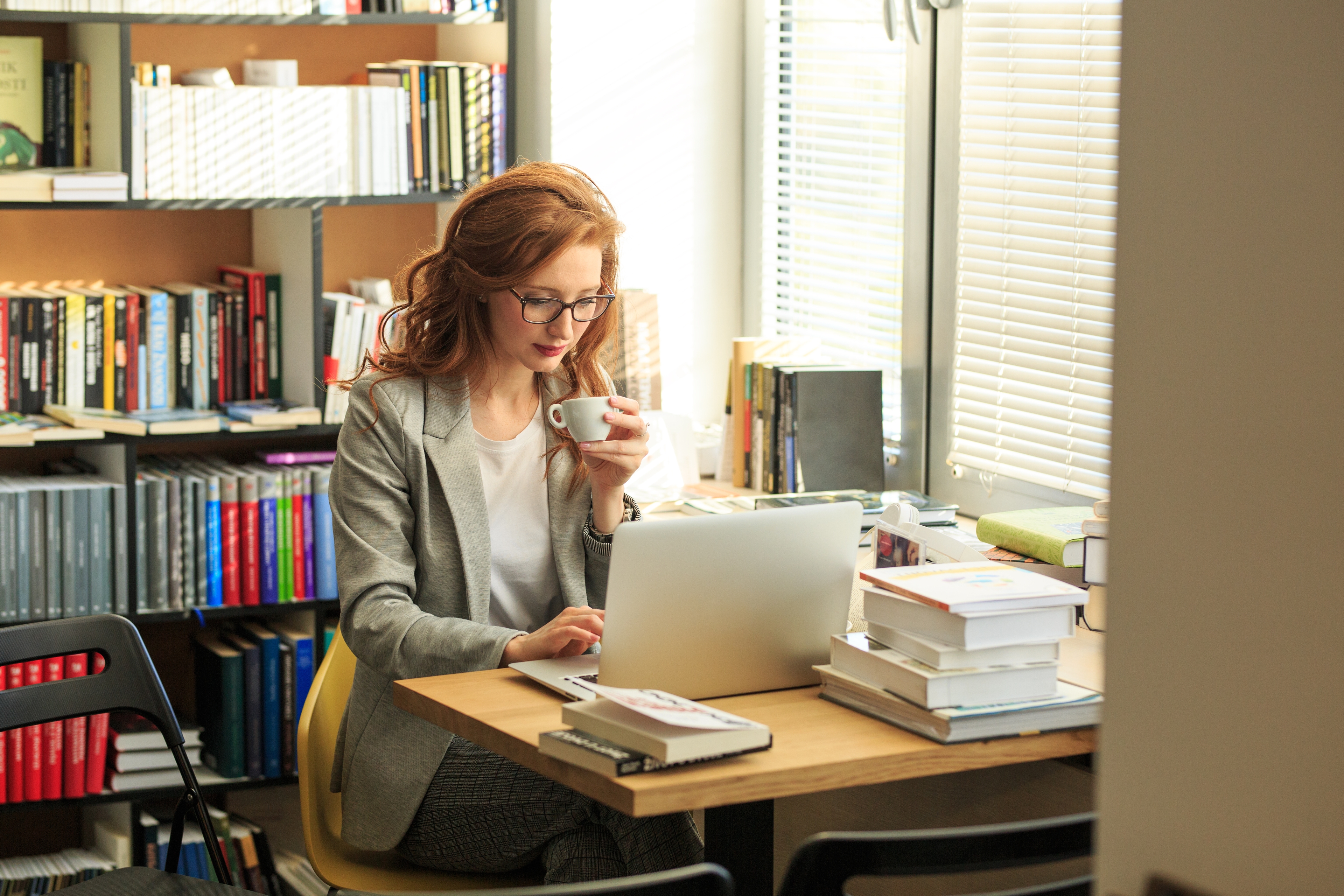 A professional woman with red hair and glasses, working on a laptop at a desk in a bright office or library, surrounded by stacks of books and holding a cup of coffee.