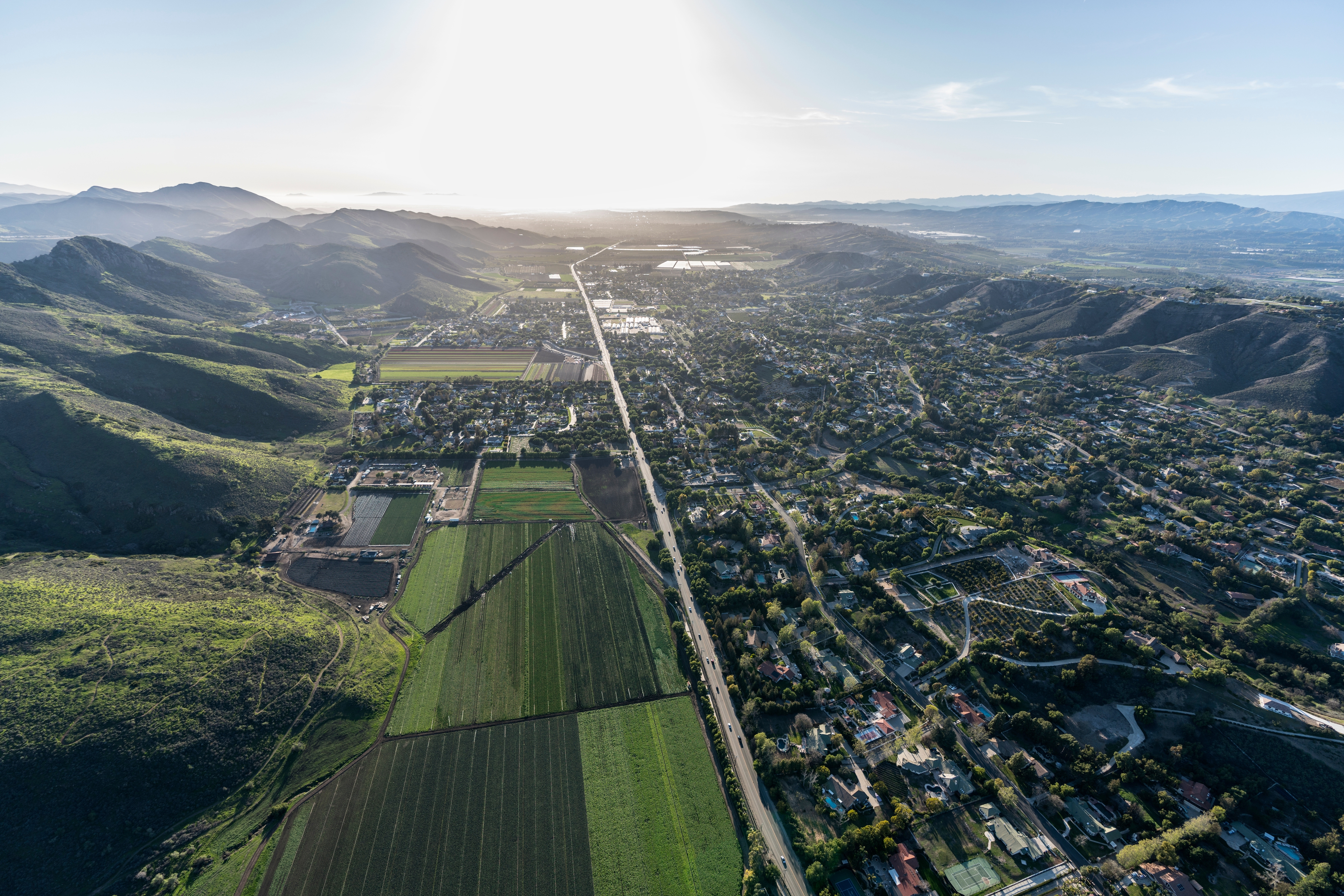 An aerial view of Santa Rosa, California, during daylight. The cityscape features a mix of residential areas and agricultural fields, bordered by rolling hills and mountains. A long road cuts through the landscape, leading toward the horizon where sunlight softly illuminates the sky. 