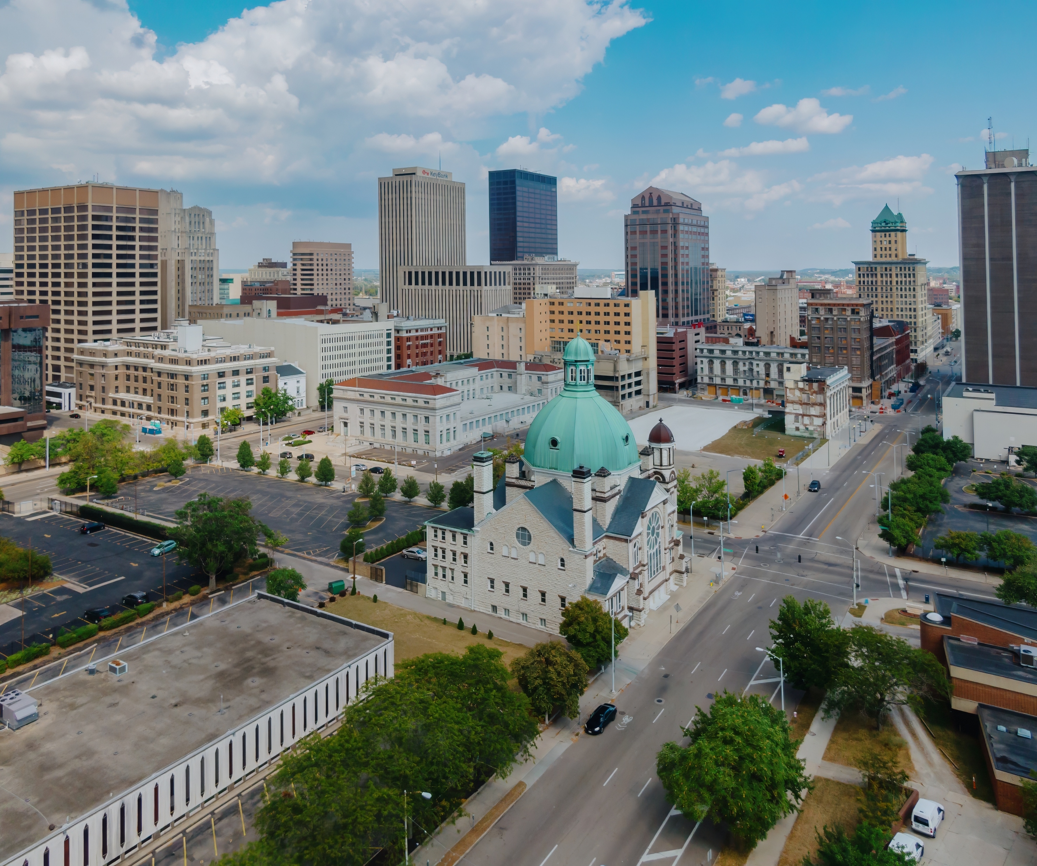 Aerial view of downtown Dayton, Ohio, highlighting the historic Sacred Heart Church with its green dome, surrounded by modern high-rises and tree-lined streets under a partly cloudy sky.