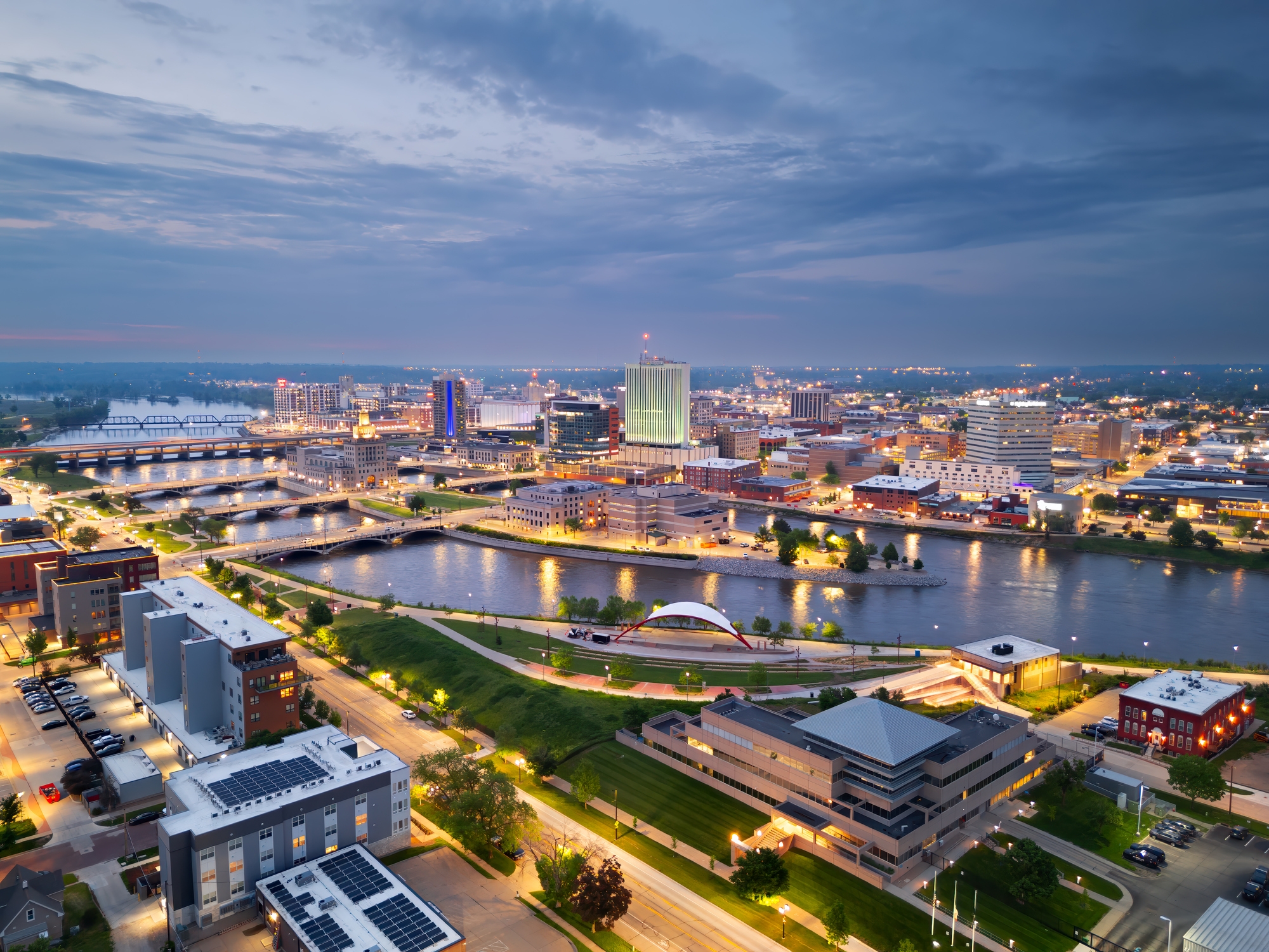 Cedar Rapids, Iowa, as seen from above during twilight, with bridges spanning the Cedar River and glowing lights highlighting the downtown area.