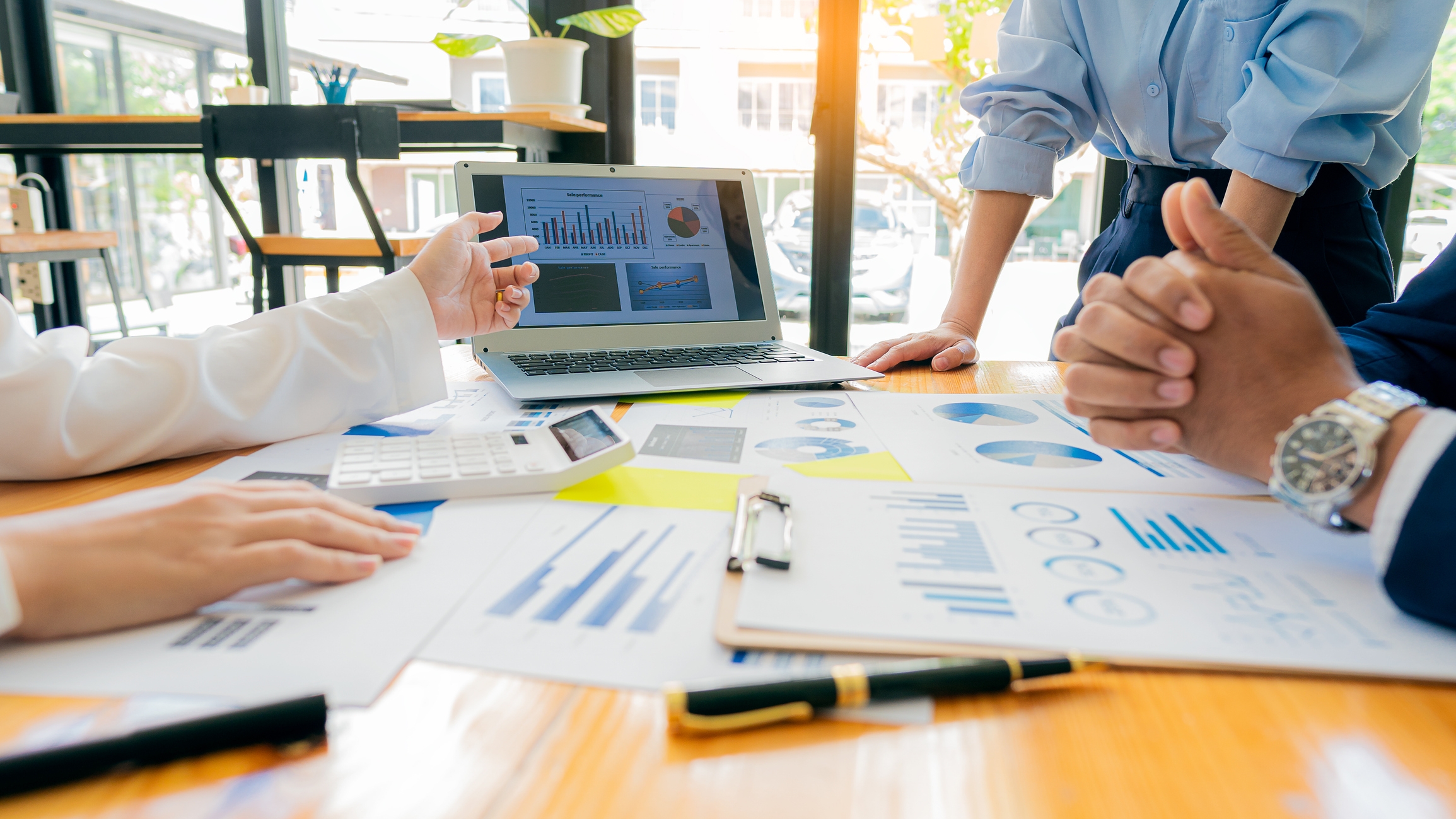 Close-up of a business meeting with financial charts and graphs on paper and a laptop screen, as colleagues discuss data analysis around a sunny office table.