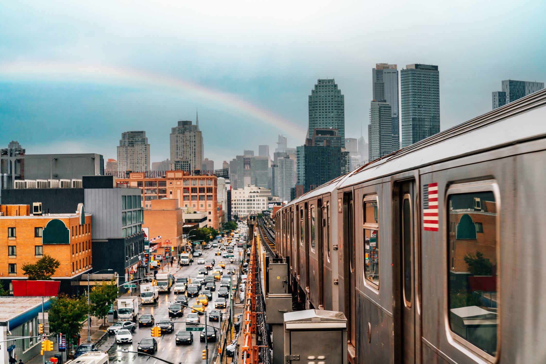 An urban scene in Queens, New York, showing a subway train traveling on elevated tracks alongside a busy street filled with cars and yellow taxis. The backdrop features modern skyscrapers and apartment buildings under a cloudy sky, with a faint rainbow arching above the cityscape.