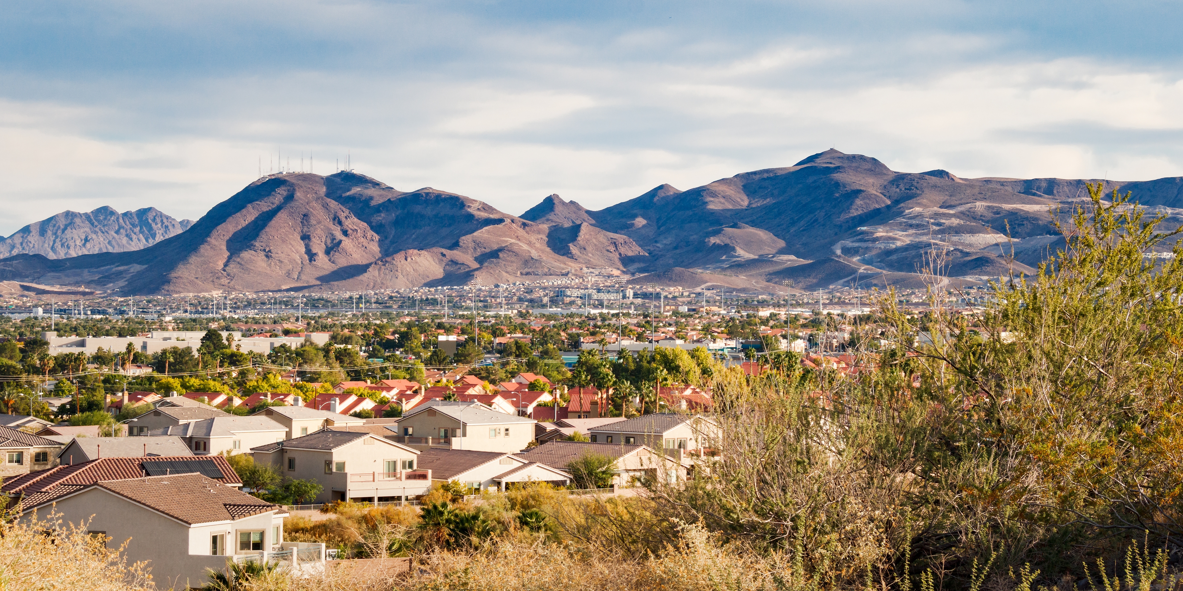 View of Henderson, Nevada with suburban homes in the foreground and rugged desert mountains rising in the background under a partly cloudy sky.